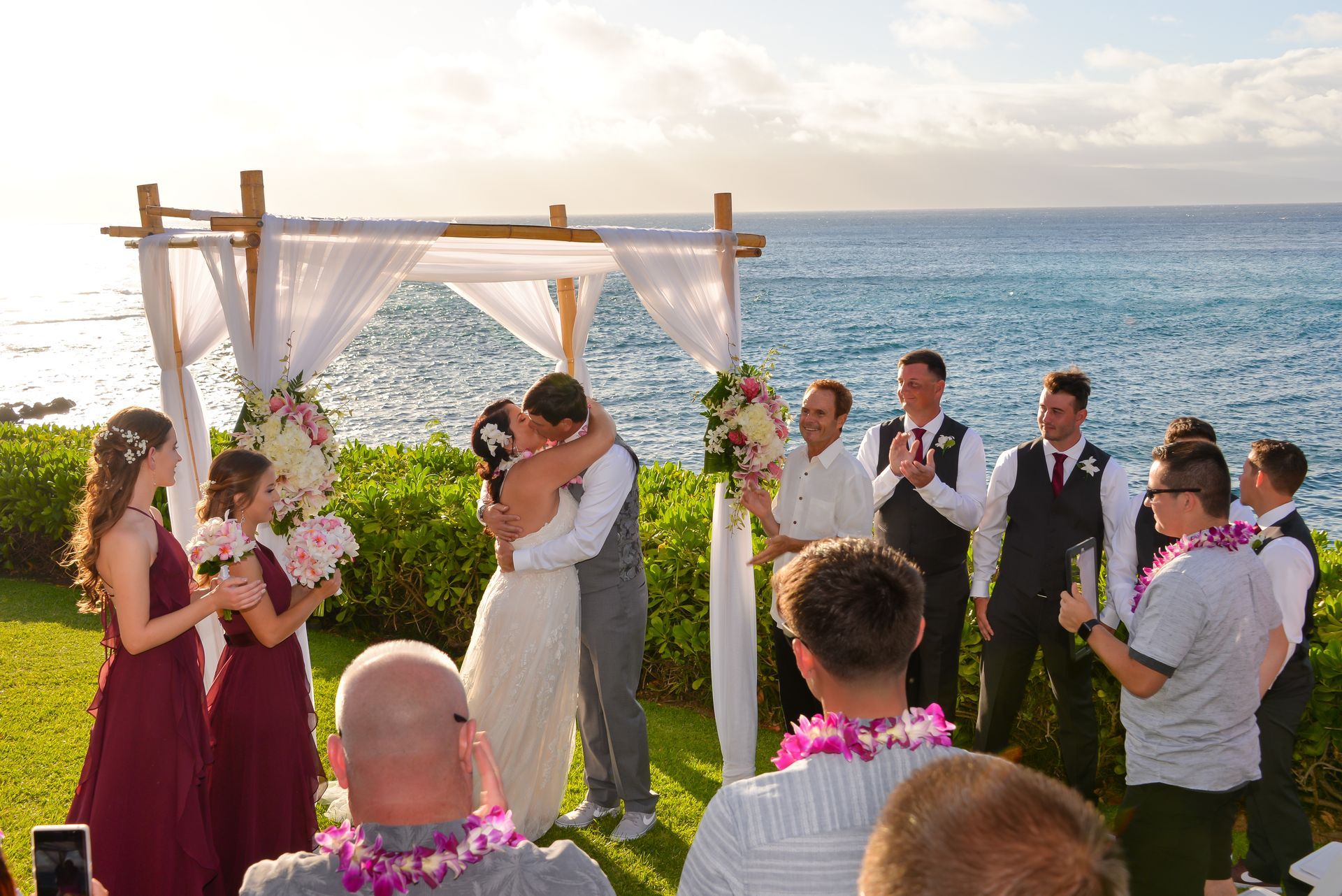 Wedding ceremony by the ocean; couple kisses under a draped arch, surrounded by wedding party and guests.