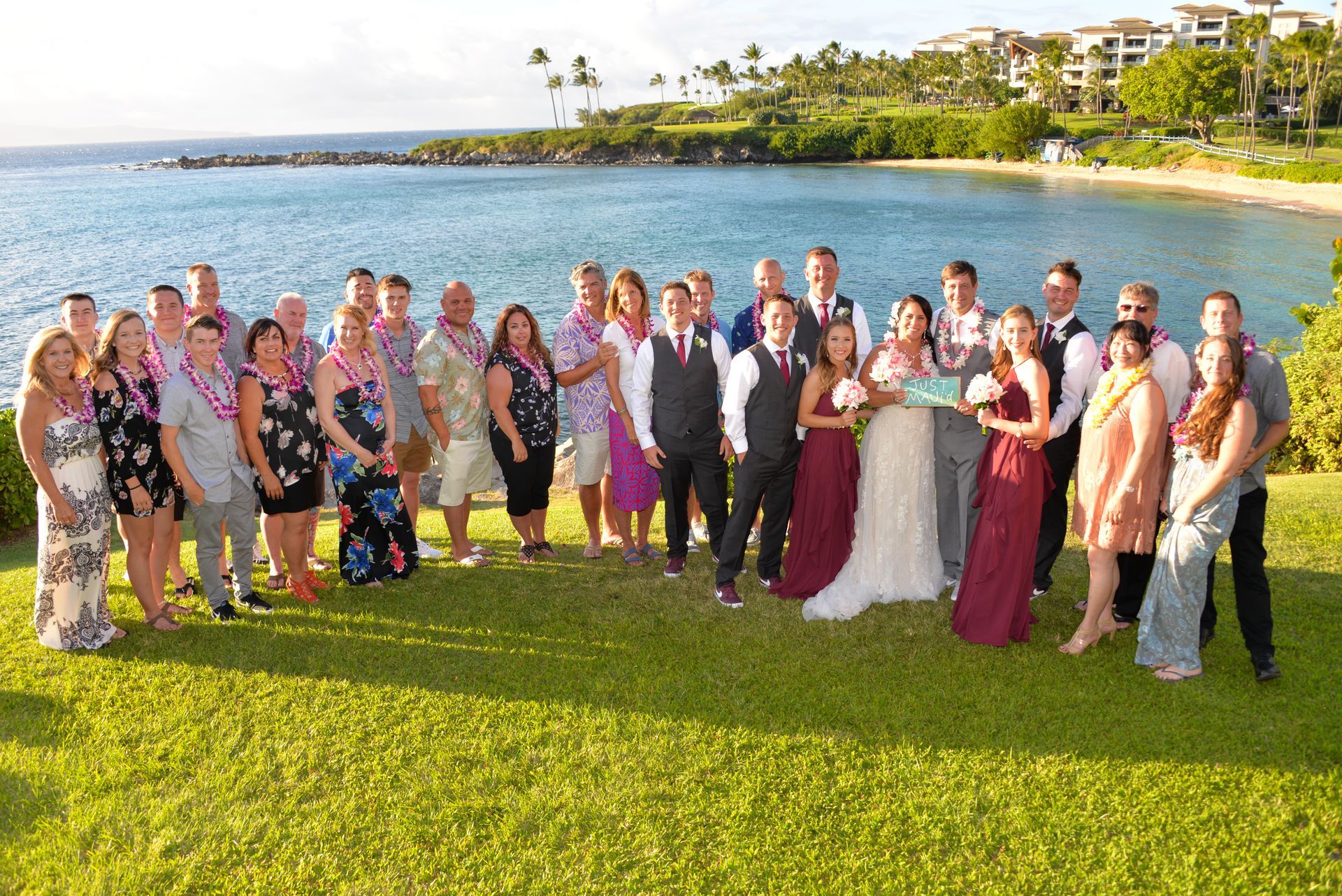 Wedding party on a grassy hill overlooking a bay. Bride and groom in center, surrounded by guests, wearing leis.