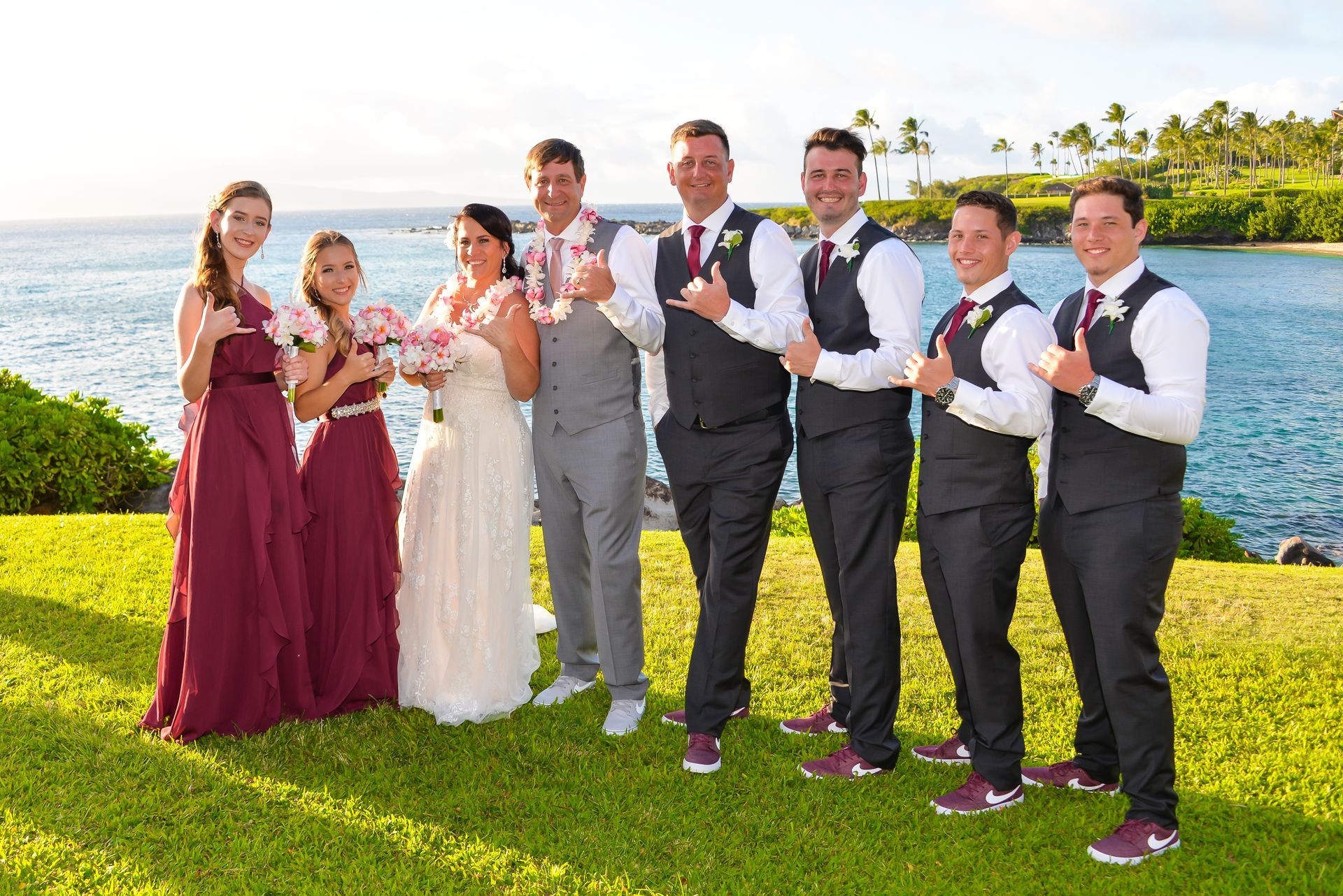 Wedding party posing on a grassy hill overlooking the ocean. Bridesmaids in burgundy dresses, groomsmen in vests.