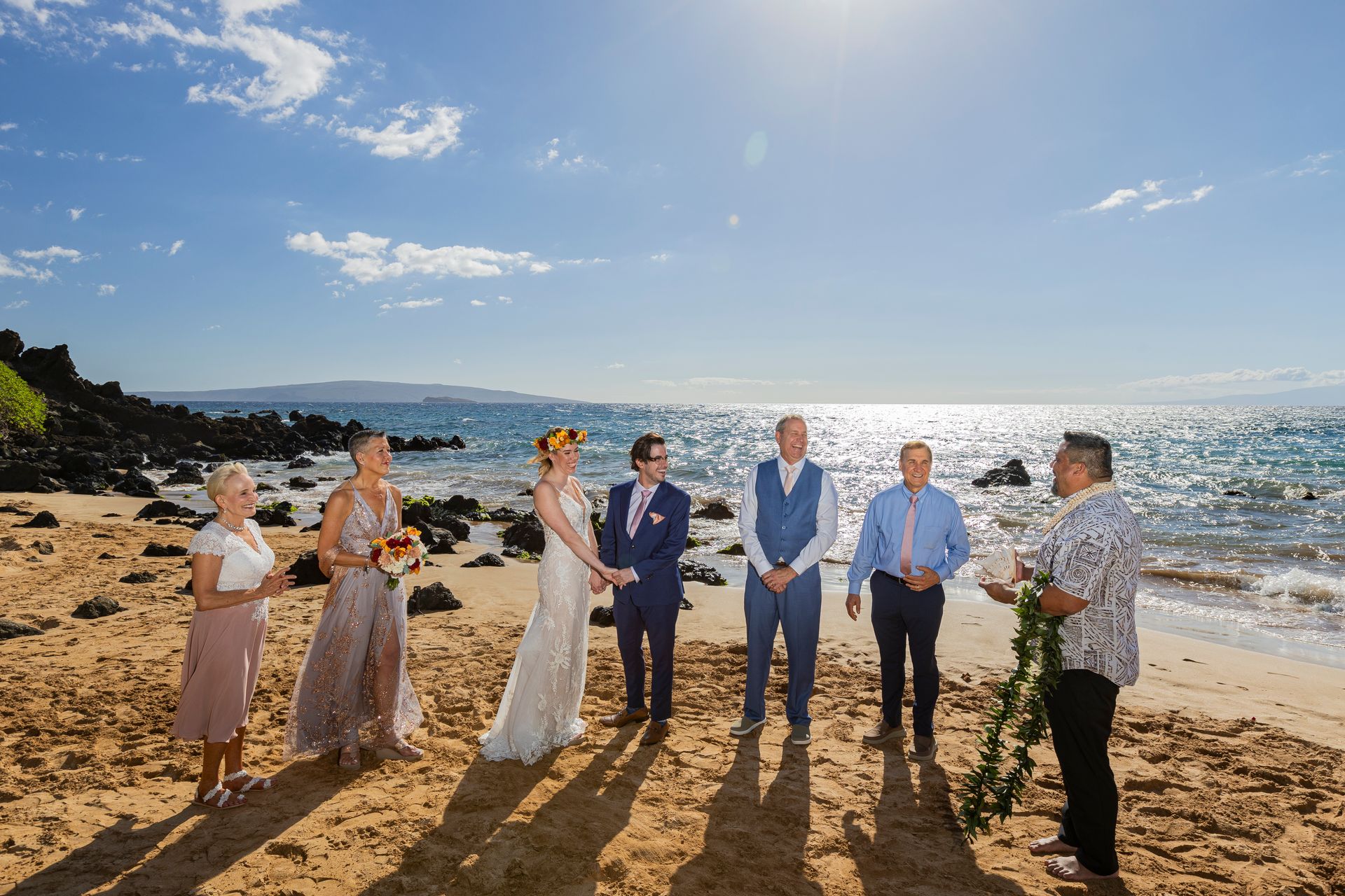 Wedding party on a sunny beach; bride, groom, officiant, and guests.