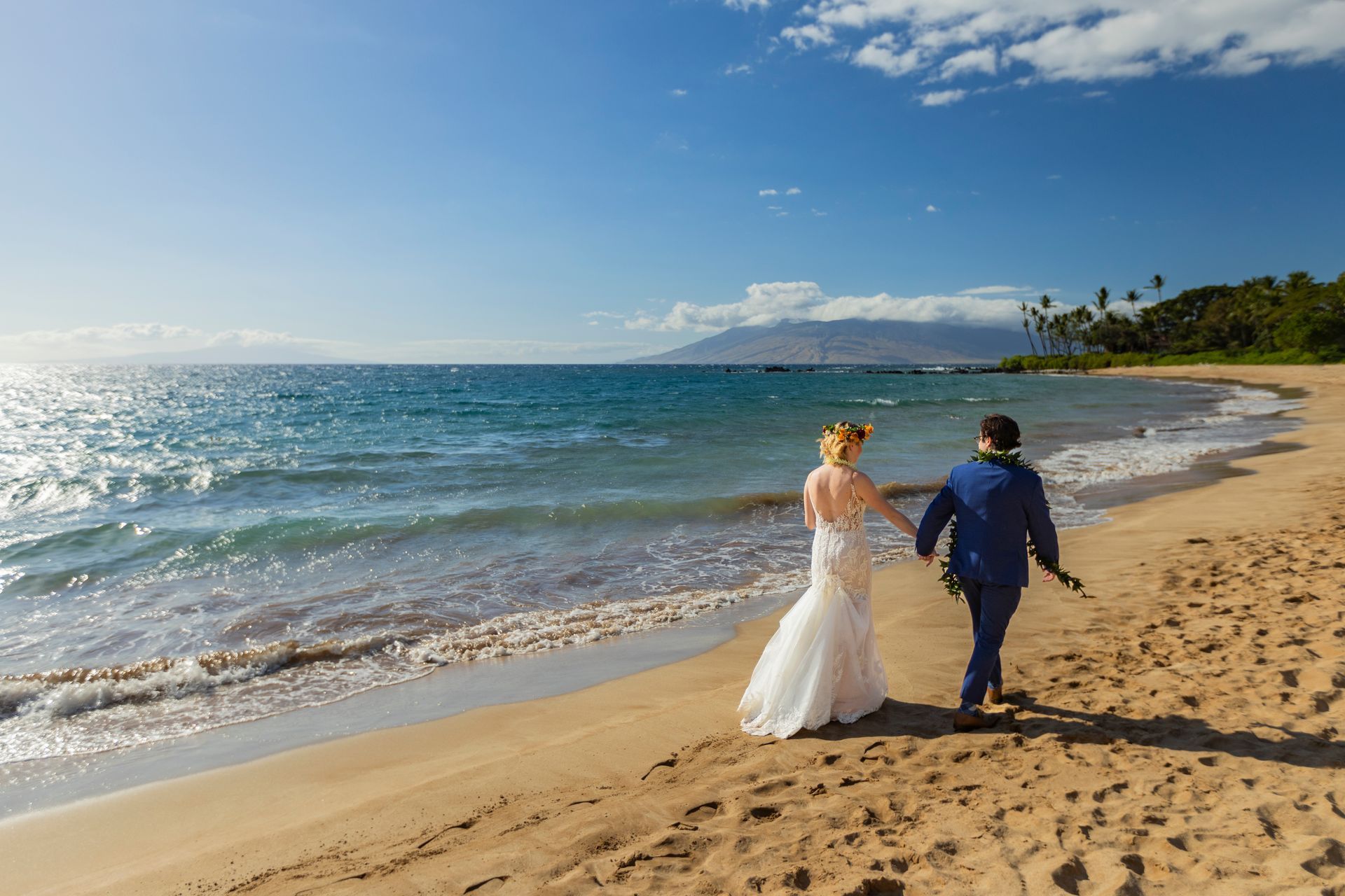 Bride and groom walk hand-in-hand on a sunny beach, ocean waves lapping, mountain in background.