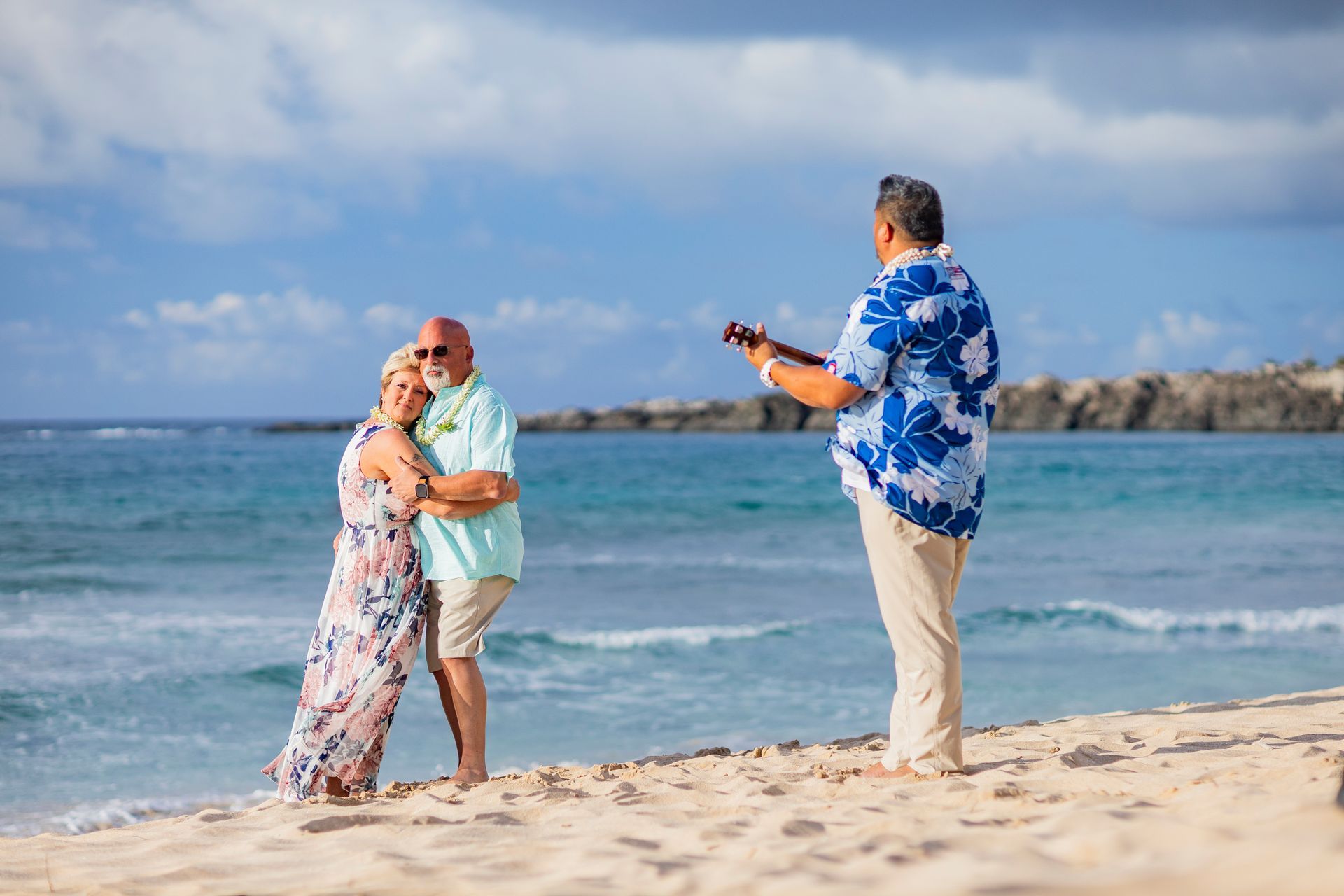 Couple on a beach, embracing while a musician in a blue shirt plays the ukulele. Blue sky and water.