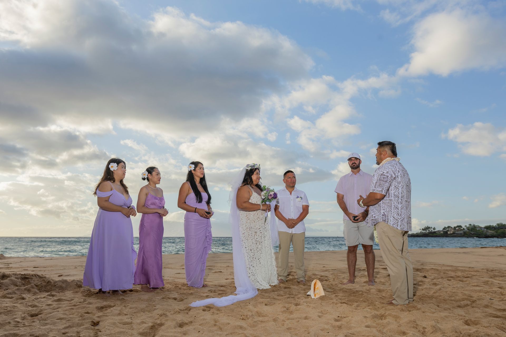 Wedding ceremony on a beach: Bride, bridesmaids in lilac, groom, officiant, guests, ocean, cloudy sky.