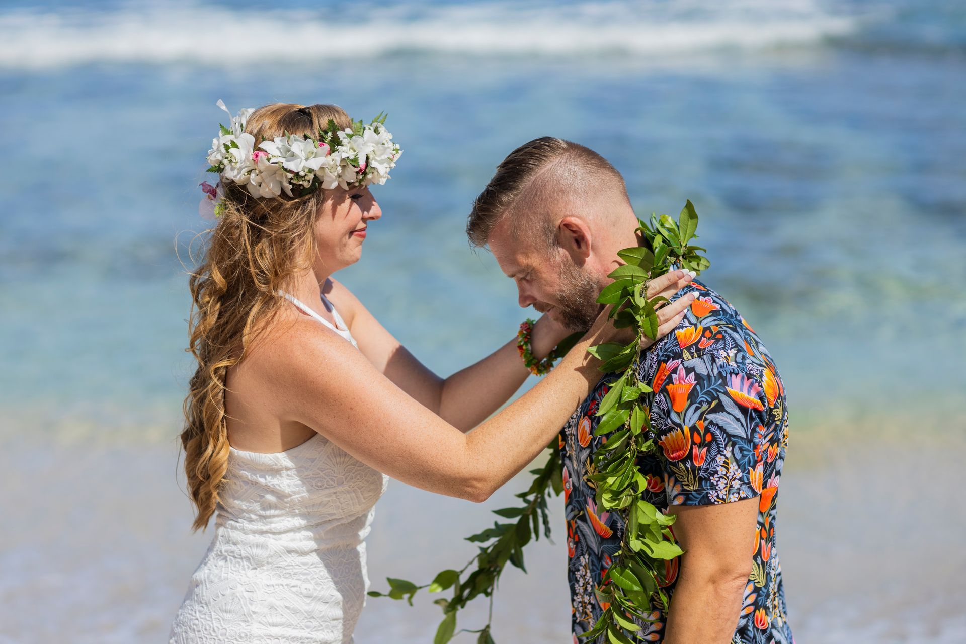 Bride places a green lei on groom during a beach wedding ceremony.