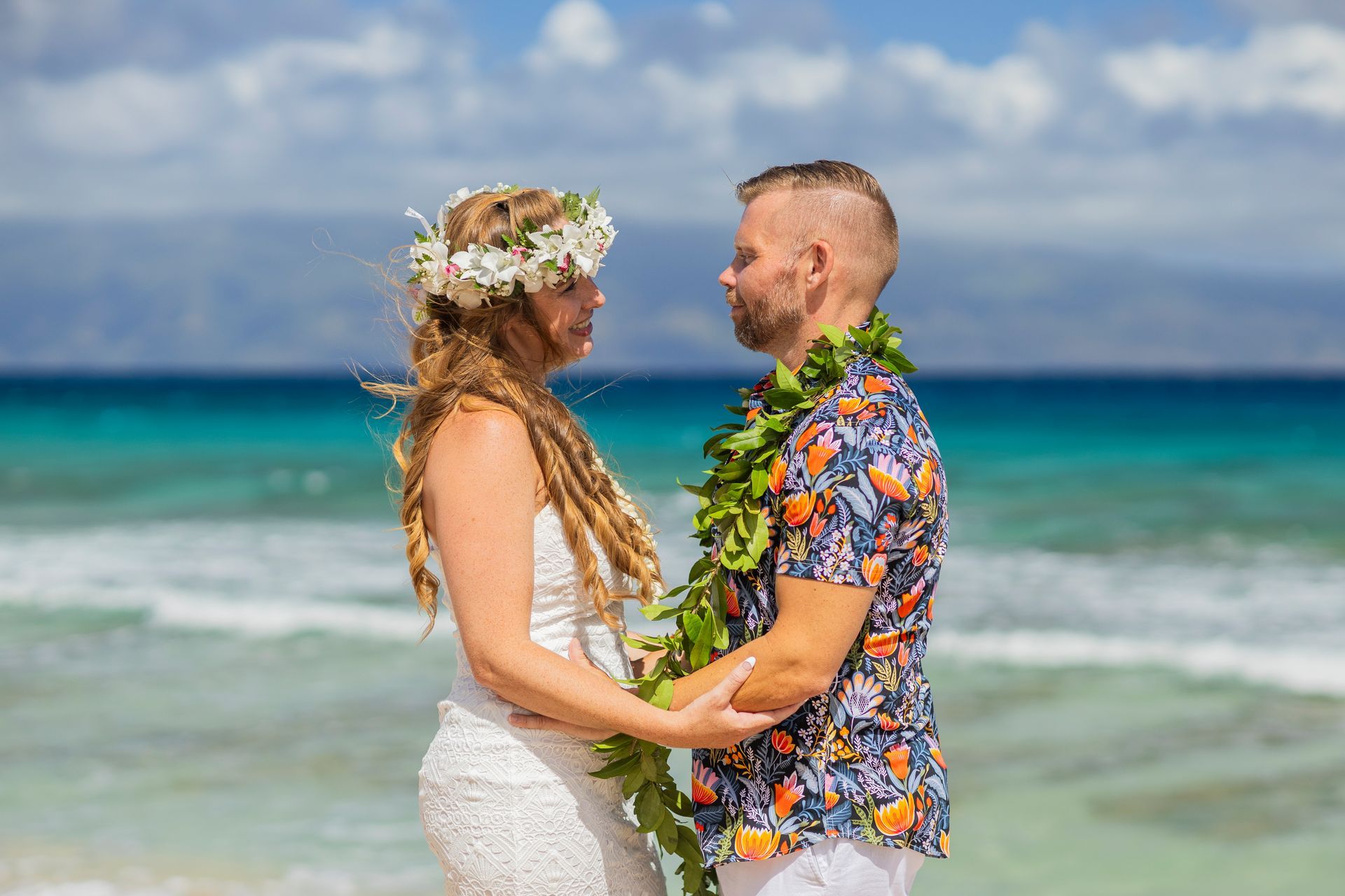 Couple holding hands on a beach, looking at each other. Bride with flower crown; groom in floral shirt. Ocean backdrop.