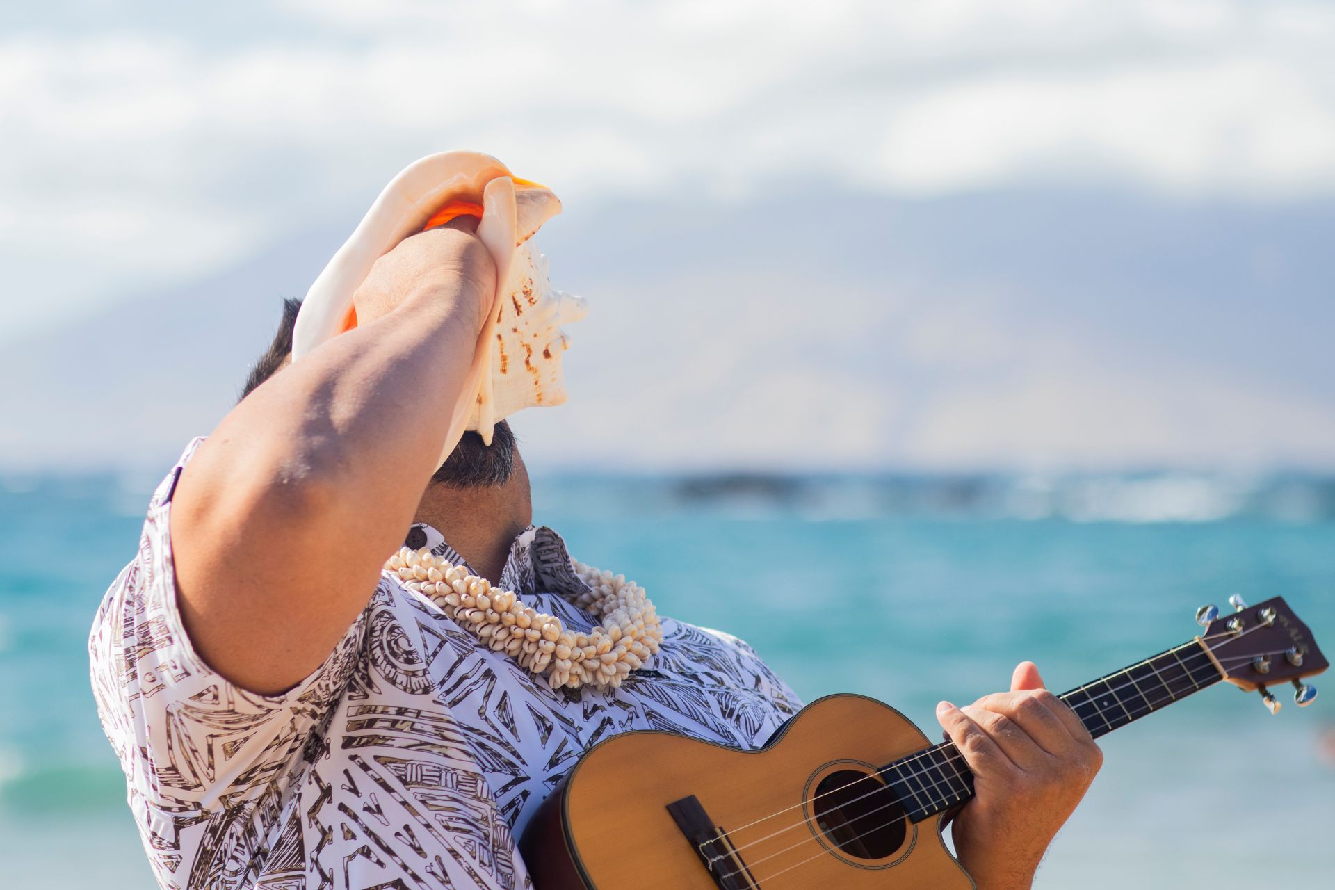 Man on beach playing ukulele, blowing conch shell, wearing lei, ocean background.