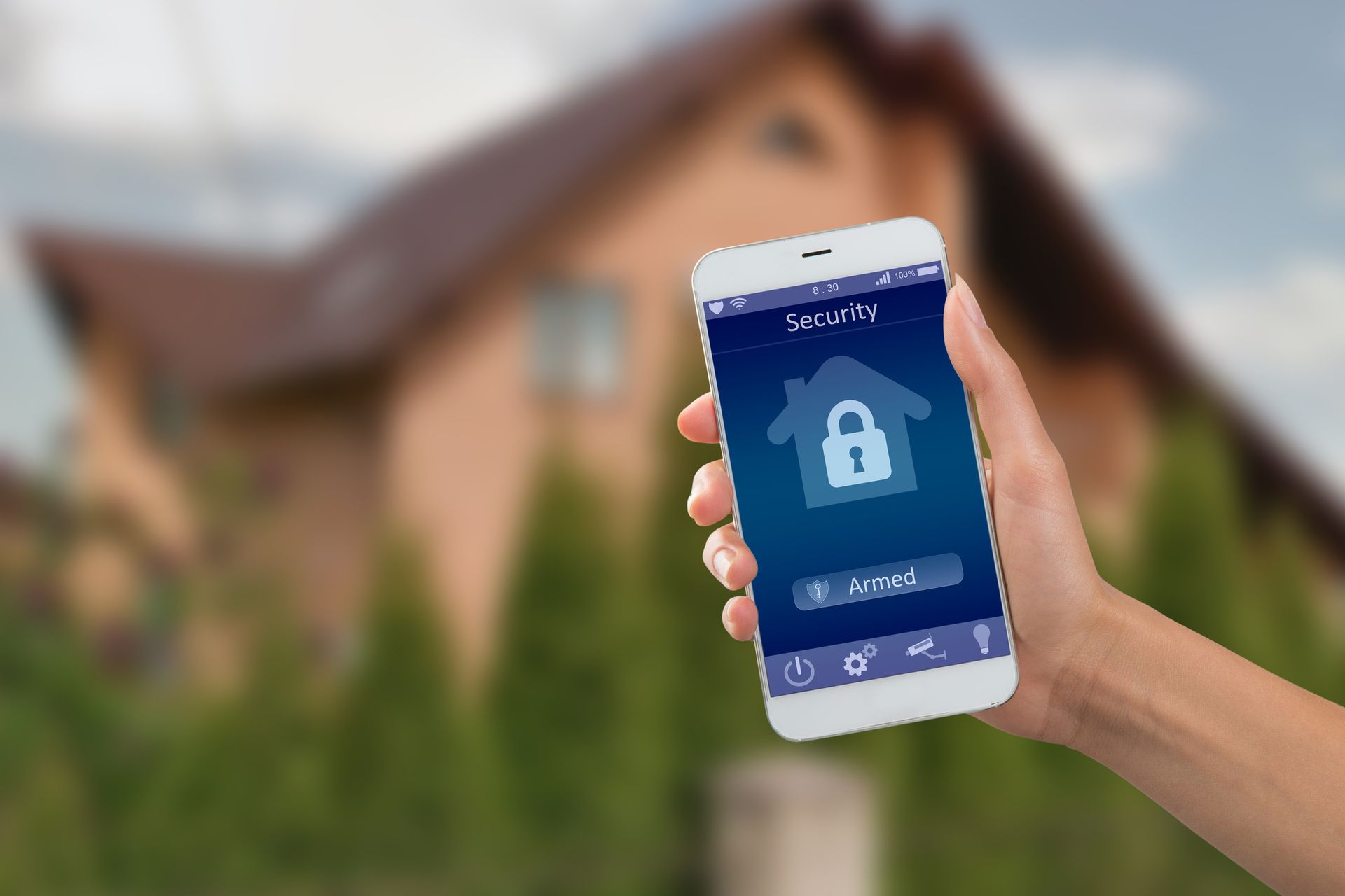 Hand holding a smartphone with a home security app interface, blurred house in background.