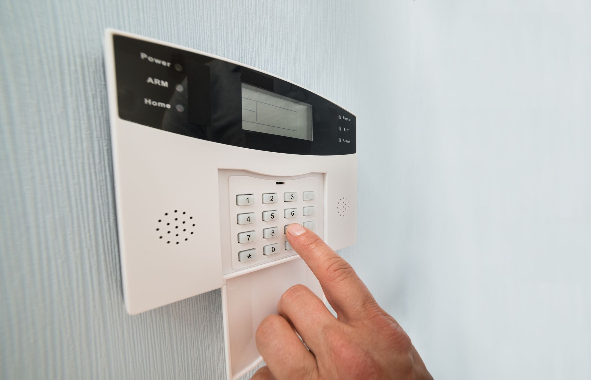 Hand pressing a security system keypad mounted on a light blue wall.