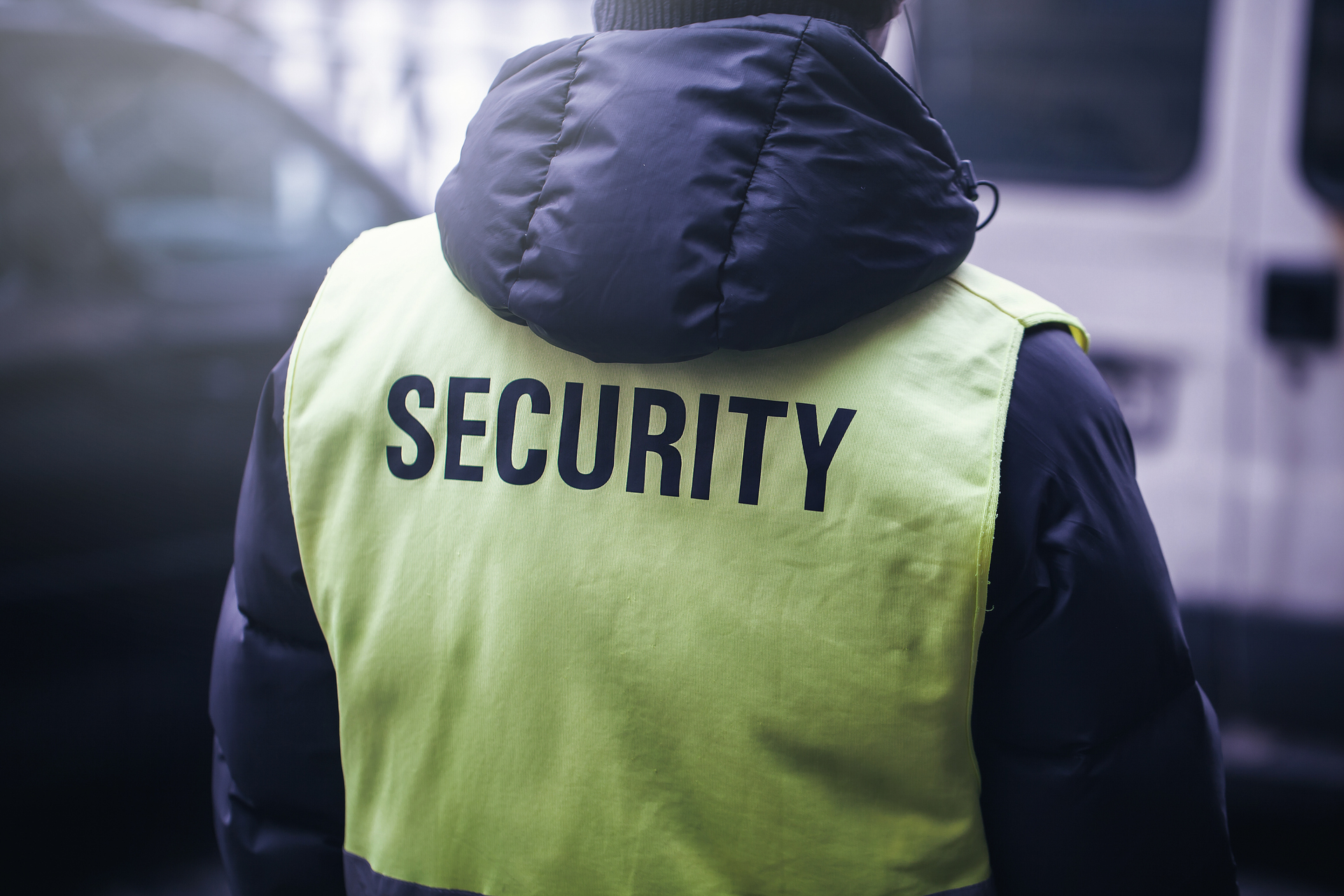 Security guard in yellow vest with "SECURITY" text, standing in front of vehicles.