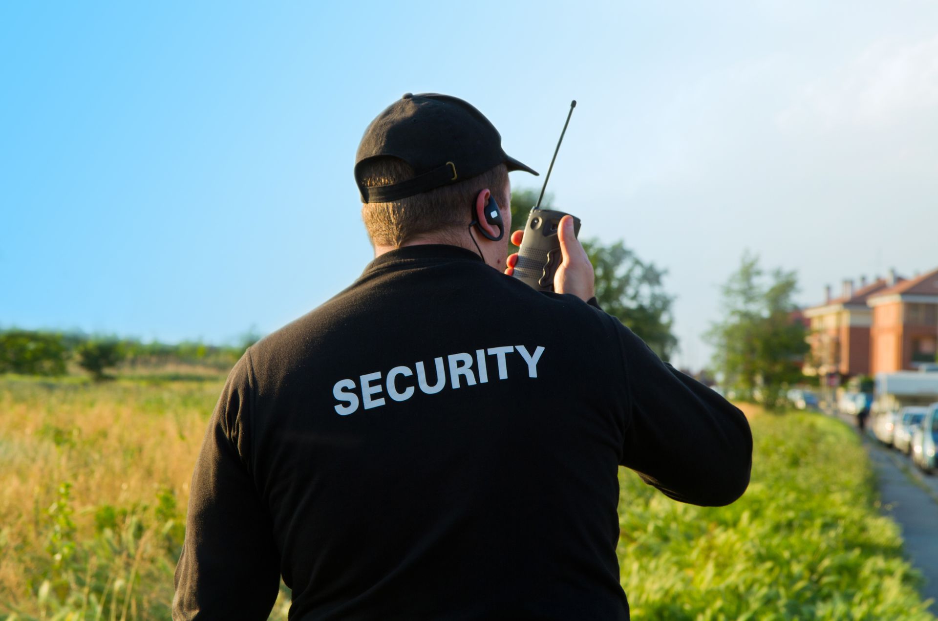 Security guard wearing black uniform talking on a radio, standing outside near a field and buildings.