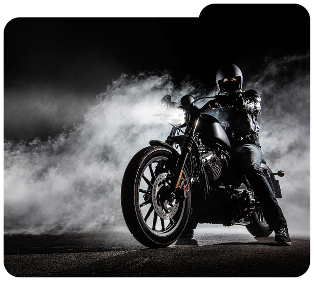 Motorcyclist on a black bike in smoky darkness, headlights glowing against a dramatic backdrop