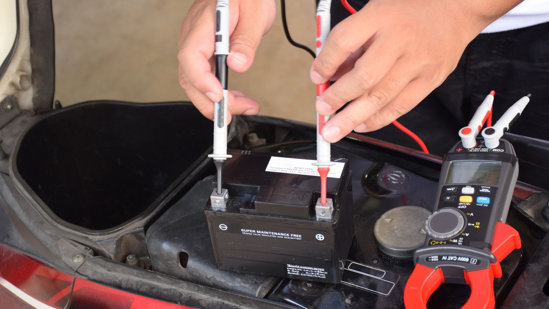Hands testing a car battery with a multimeter and probes inside the engine compartment