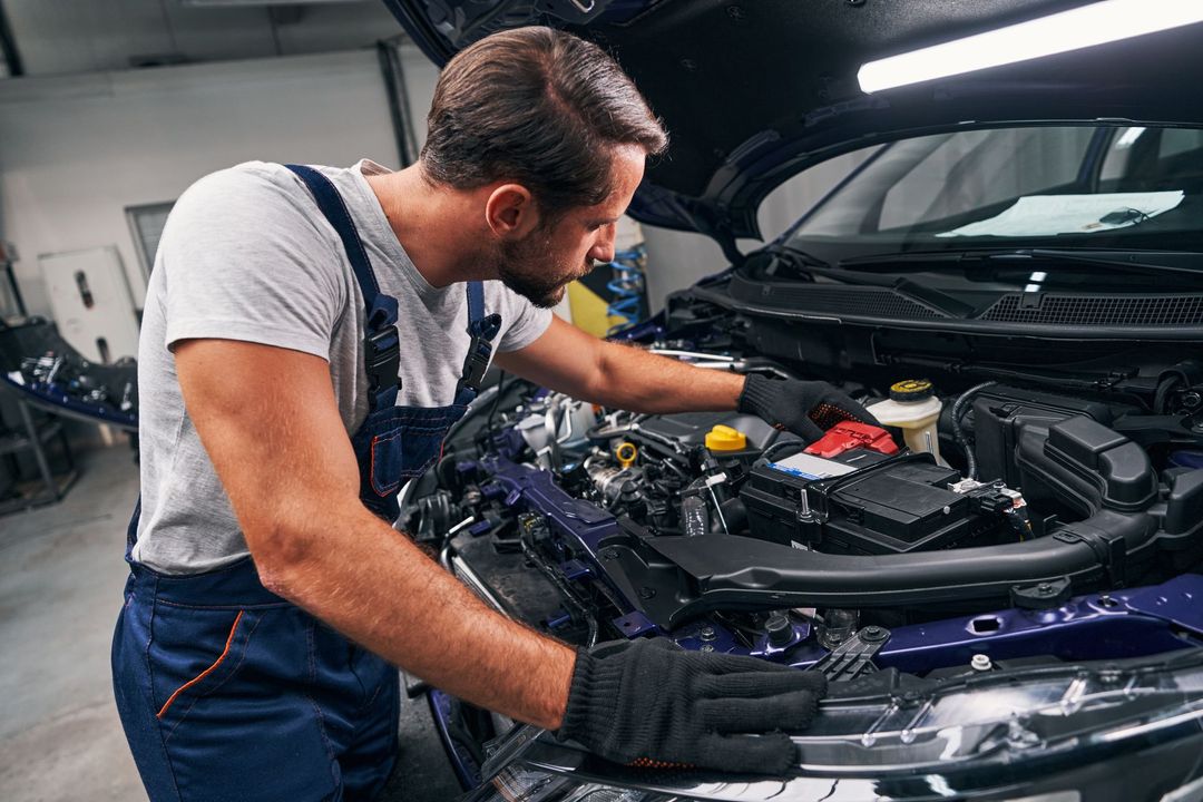 Mechanic inspecting a car engine in a garage, wearing gloves and overalls.