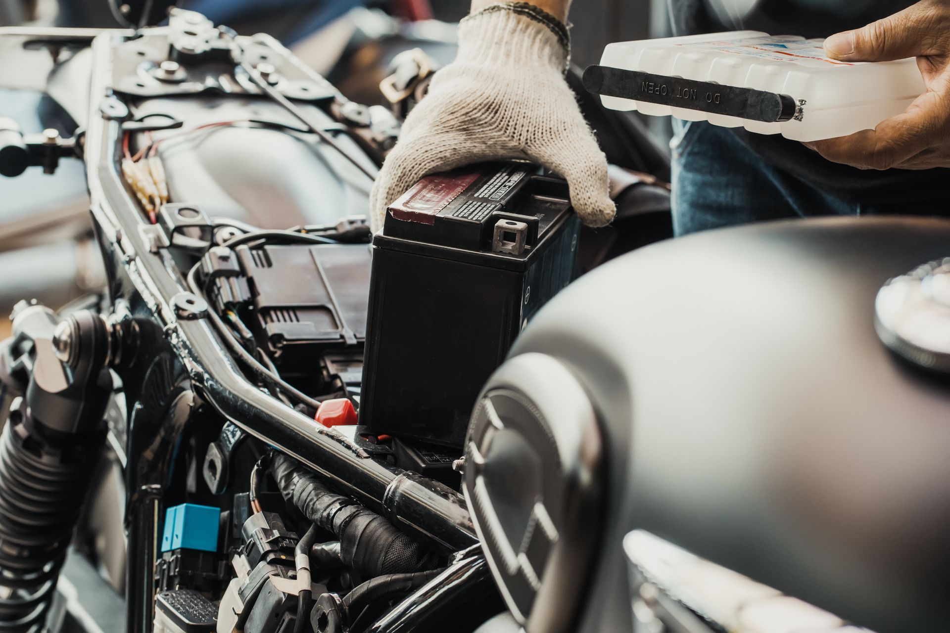 Gloved hands working on a motorcycle engine in a garage, with a black fuel tank in the foreground