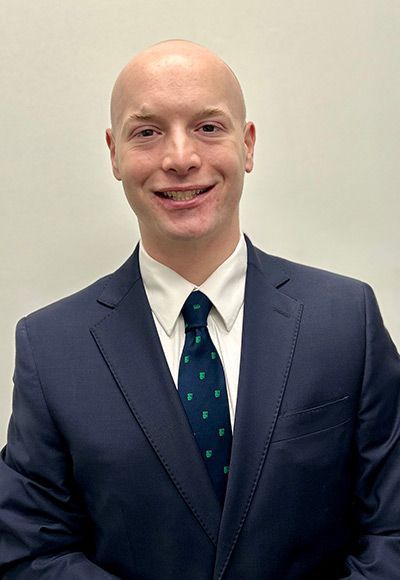 Man with a shaved head smiles, wearing a navy suit and a patterned tie, standing in front of a white wall.