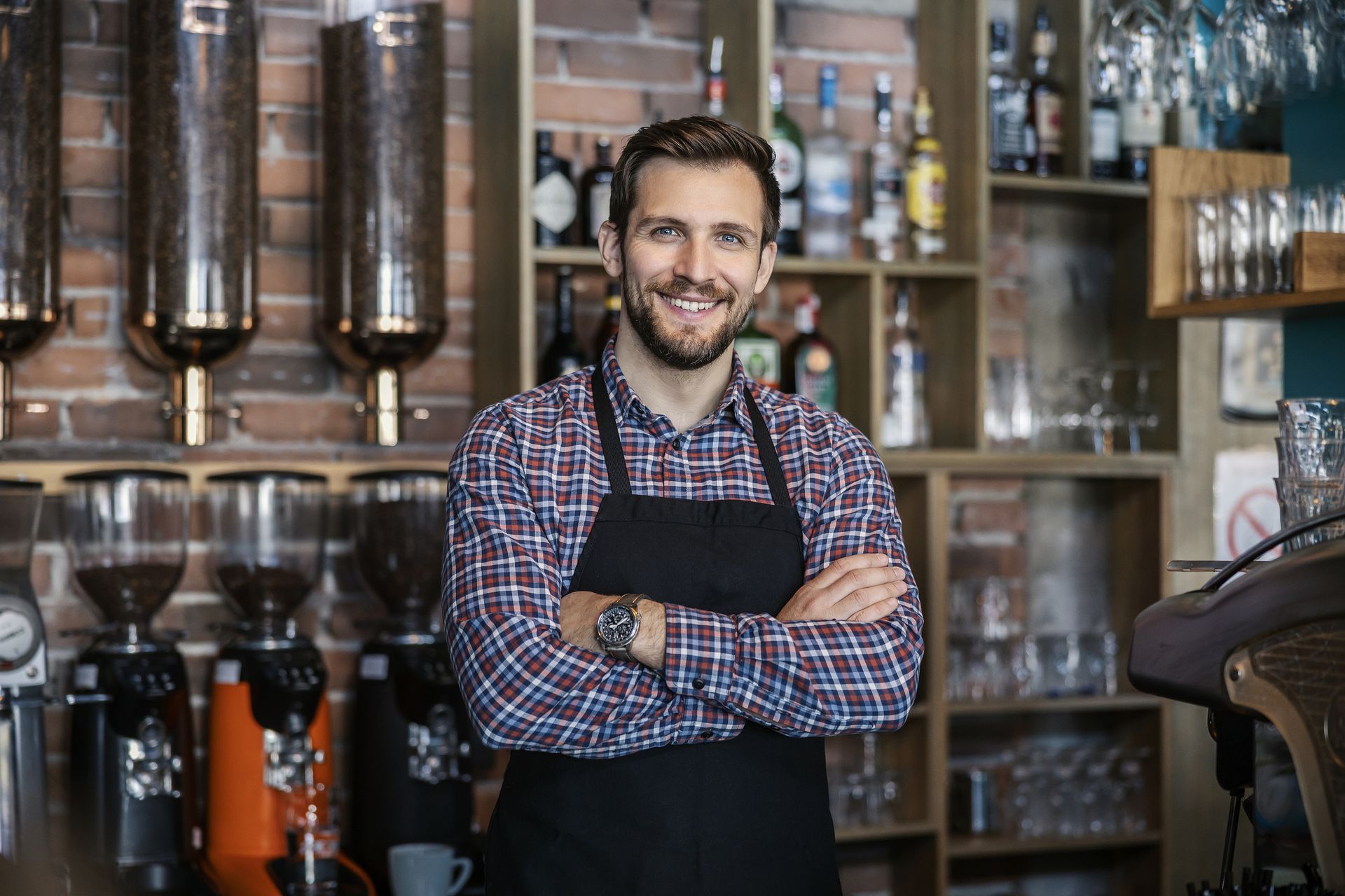 A man in an apron is standing in a coffee shop with his arms crossed.
