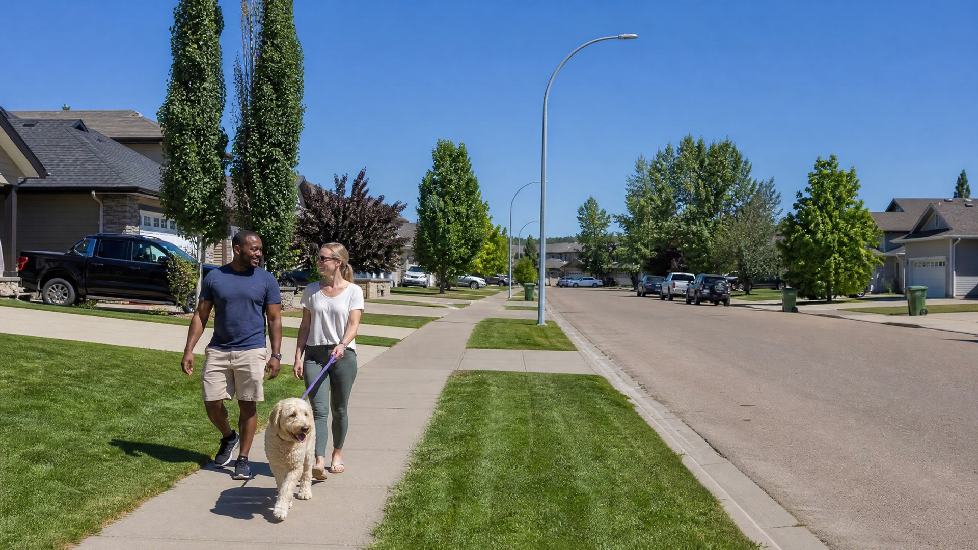 Two people walking a dog on a sunny suburban sidewalk beside a quiet street