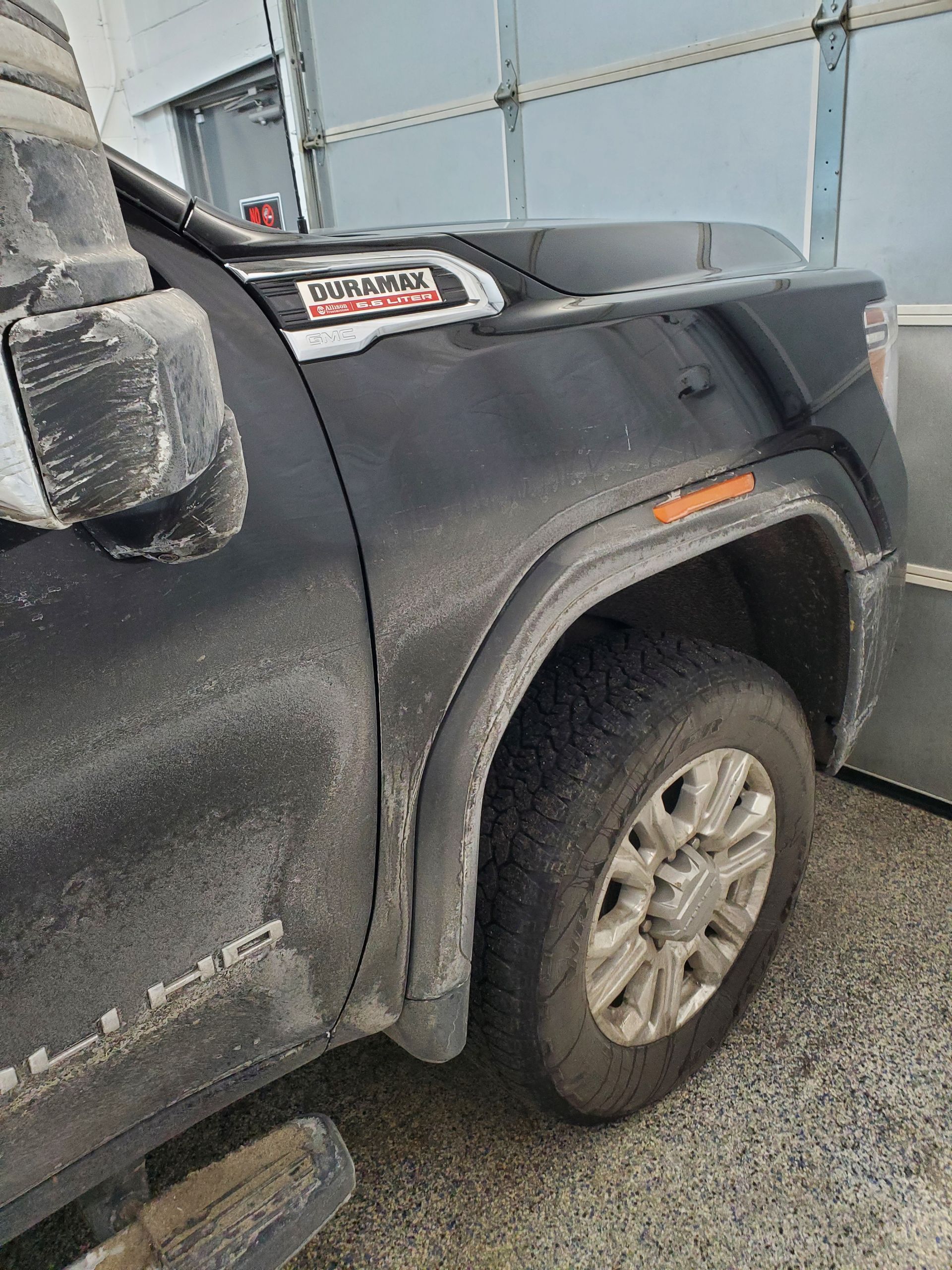 A close-up of a dirty, black GMC truck fender showing the Duramax badge, wheel, and side mirror.