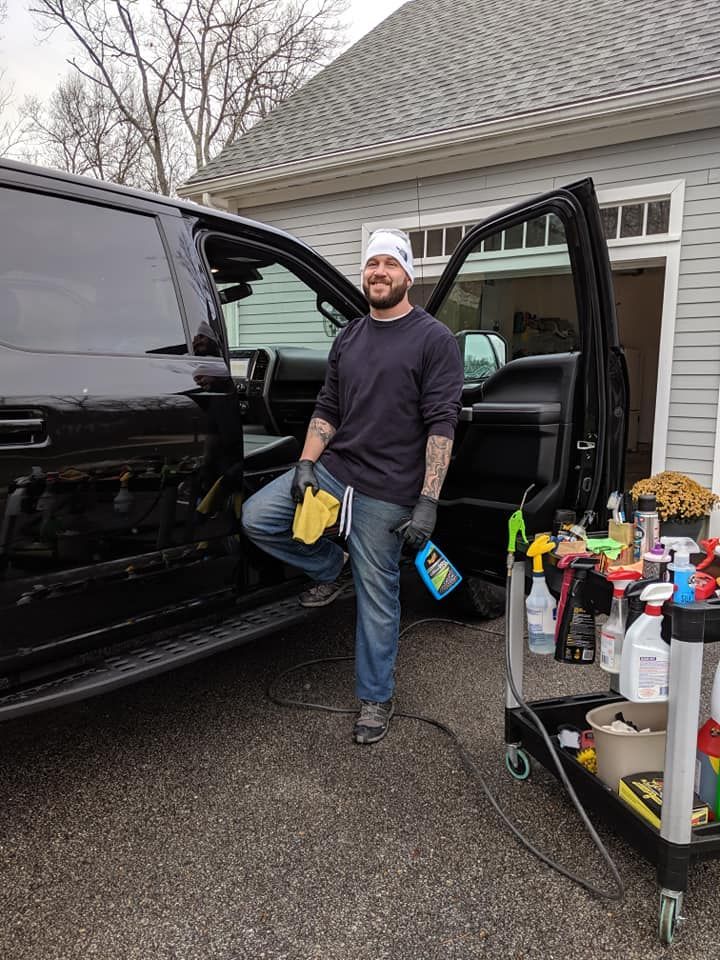 A person wearing a white beanie and gloves stands next to a black truck, holding cleaning supplies near a utility cart.