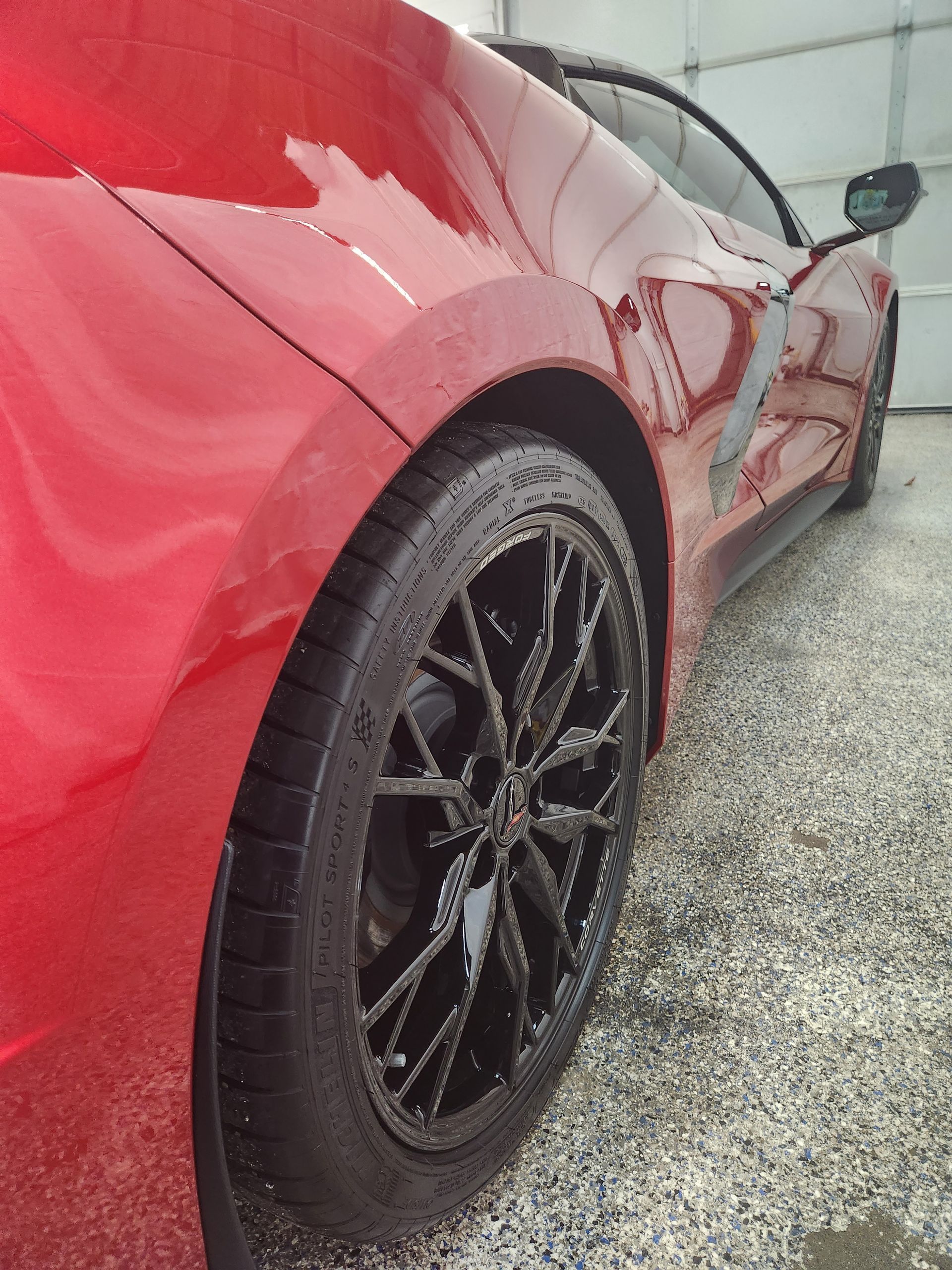 Red sports car’s rear side and black wheel on a wet gravel surface.