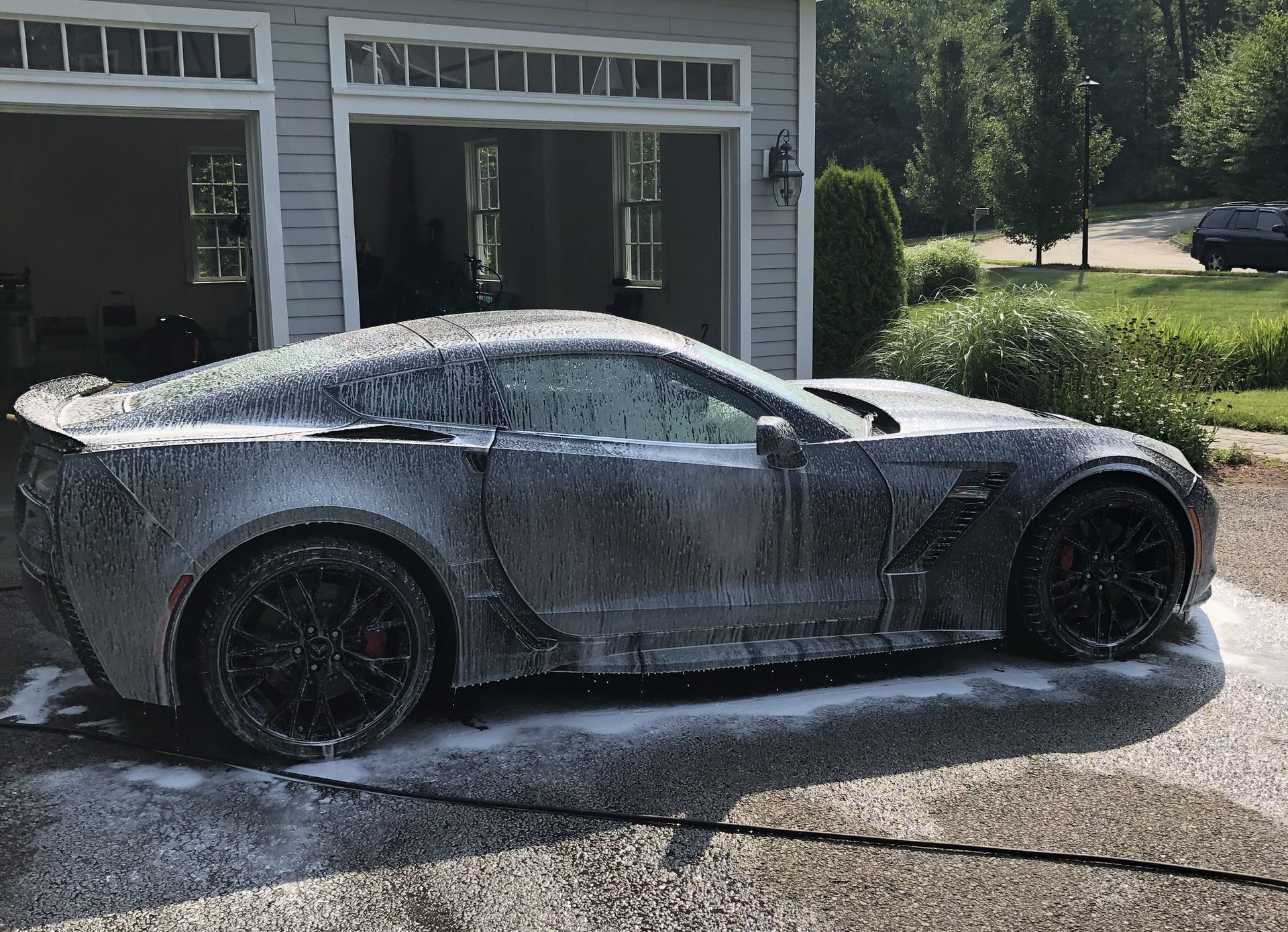 A sleek sports car covered in white cleaning foam parked on a driveway in front of a garage.