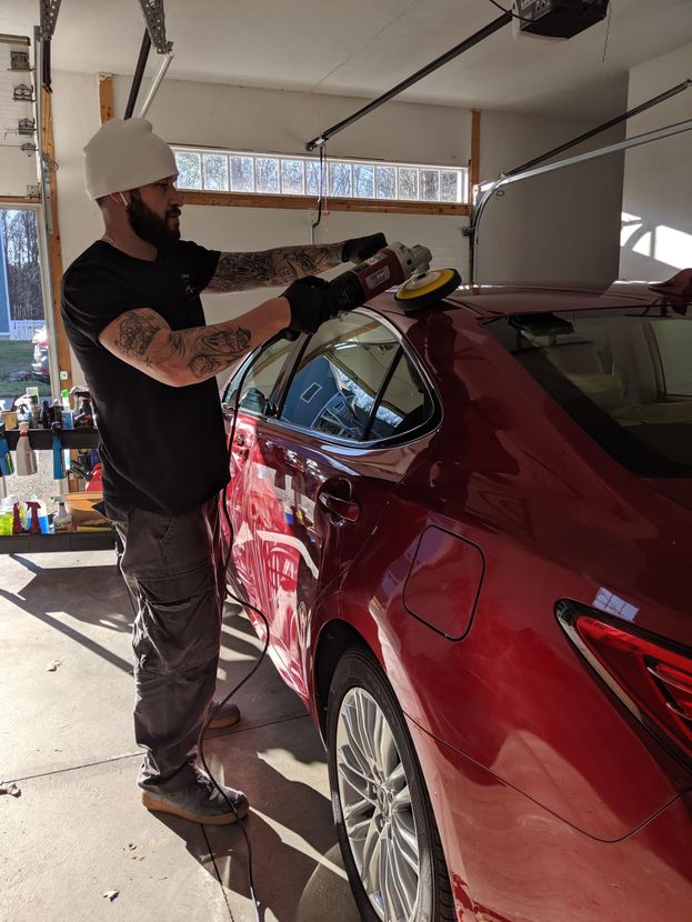 A person in a garage uses an electric polisher on the side of a red sedan.