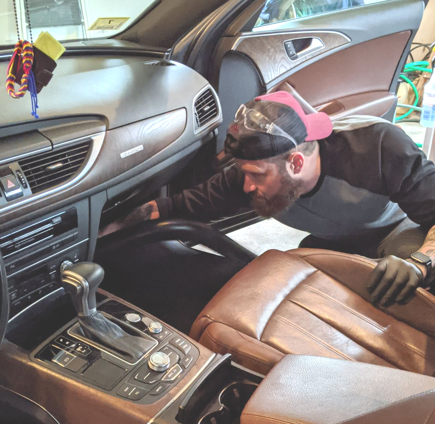 A person wearing protective eyewear and gloves uses a vacuum hose to clean the interior of a car with brown leather seats.