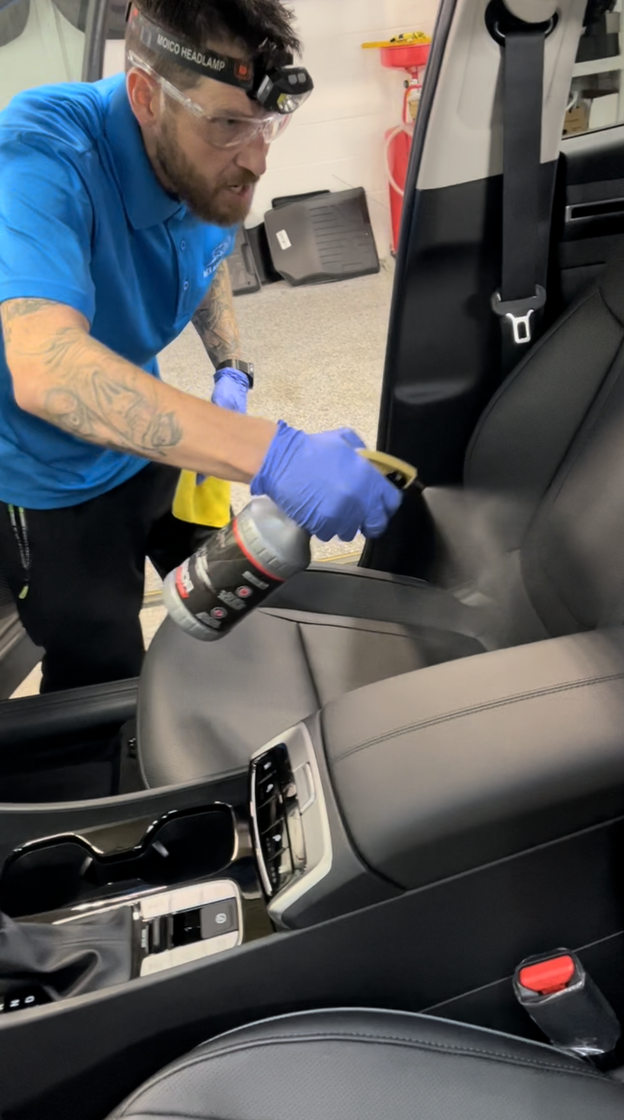 A person wearing blue gloves and safety glasses sprays a cleaning solution onto the black leather seat of a vehicle.