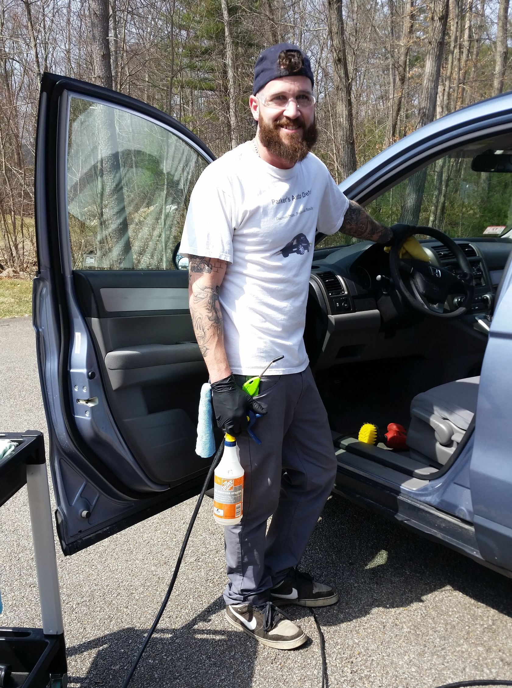 A person with a beard and tattoos stands by an open car door, holding a cleaning spray bottle and tools, outdoors.