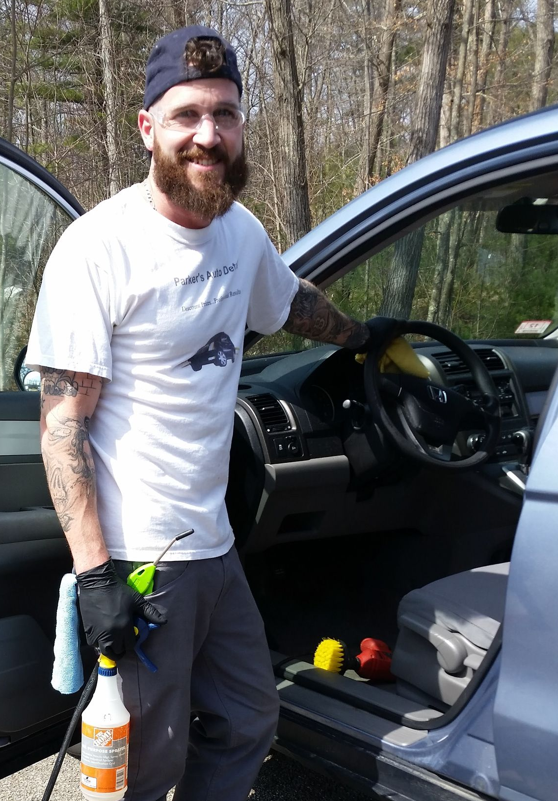 A smiling person with a beard and tattoos wearing a cap and work clothes stands by an open car door, holding a spray bottle.
