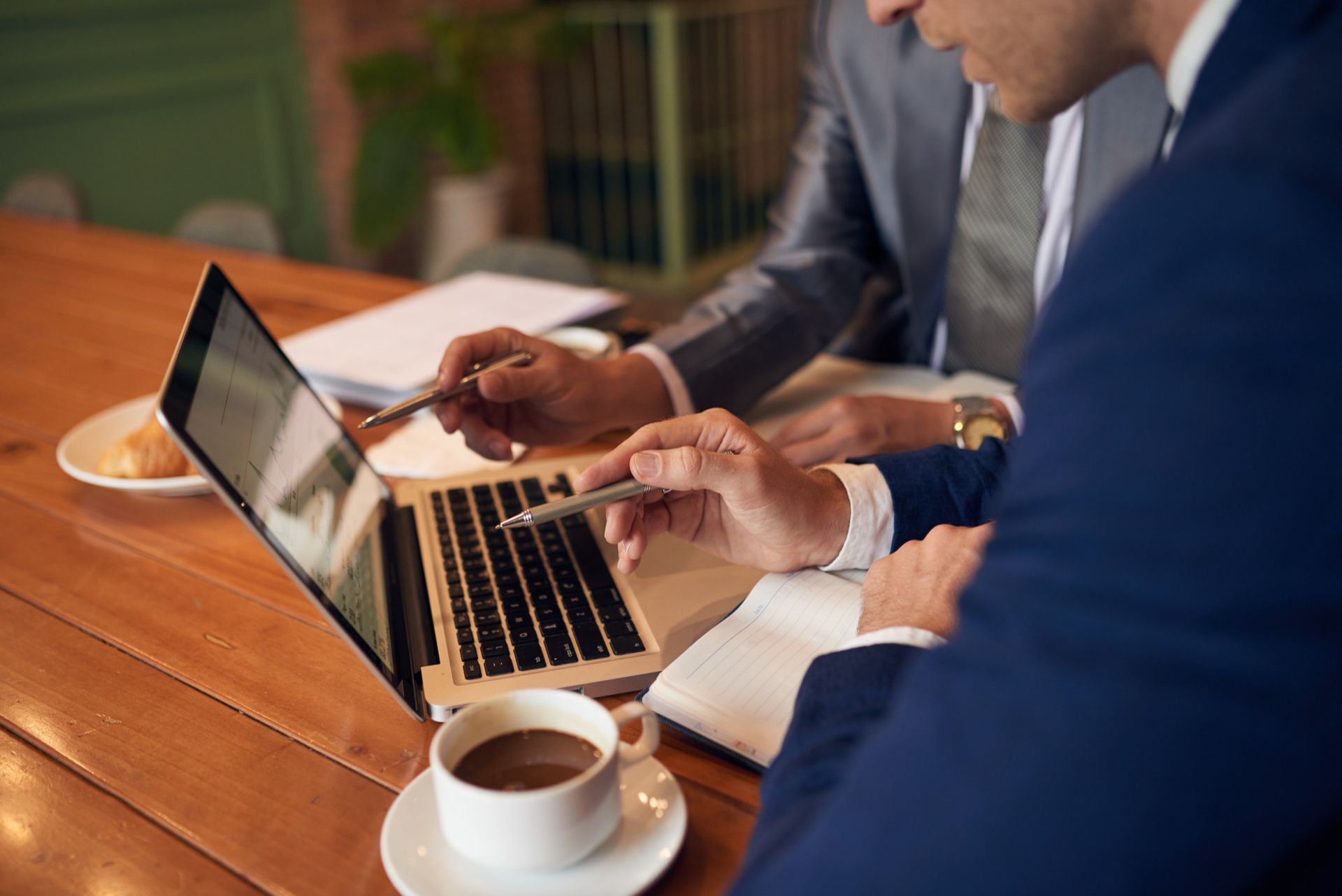 People in business attire work on a laptop at a wooden table with coffee cups.