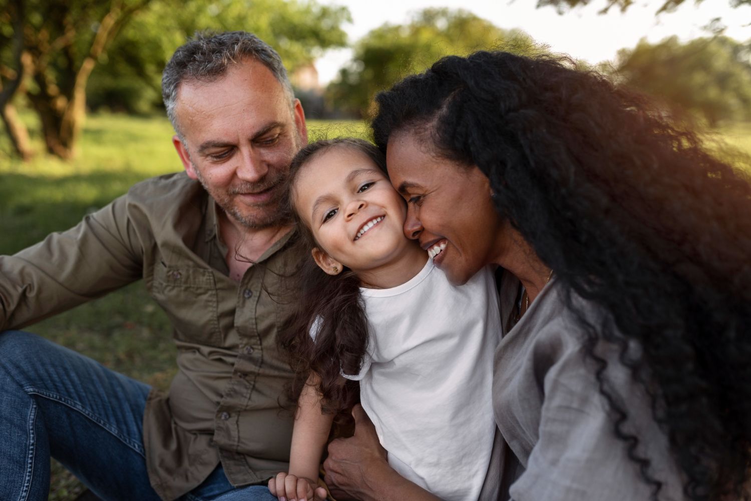 Family sitting close together outdoors, smiling and cuddling in warm sunlight.