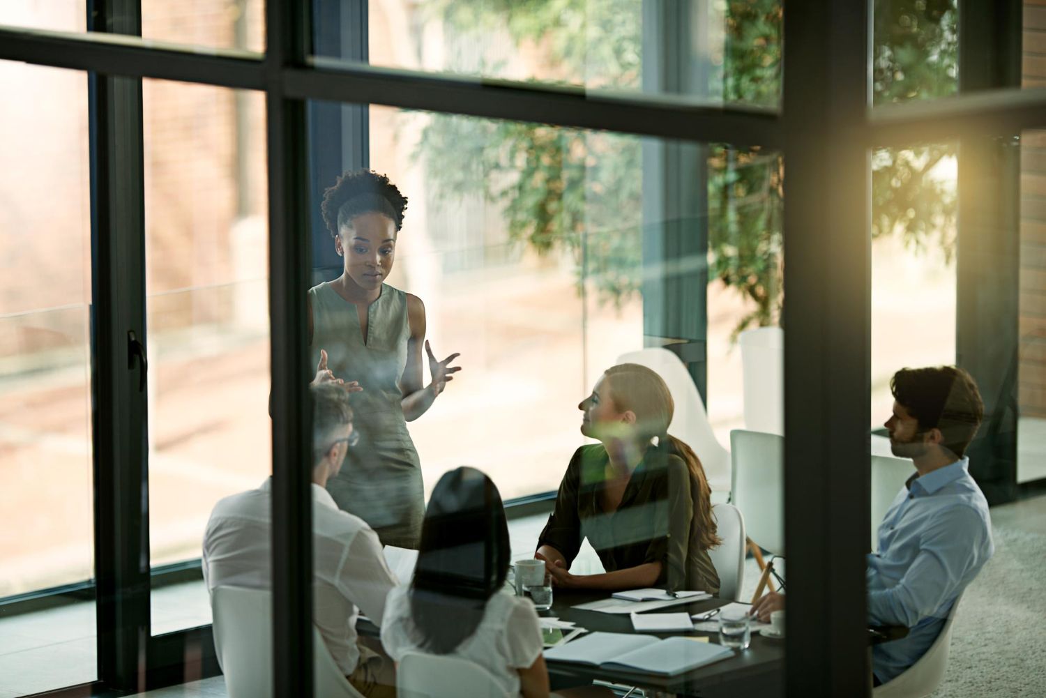 People meeting in a glass-walled conference room, with one person presenting near a whiteboard.