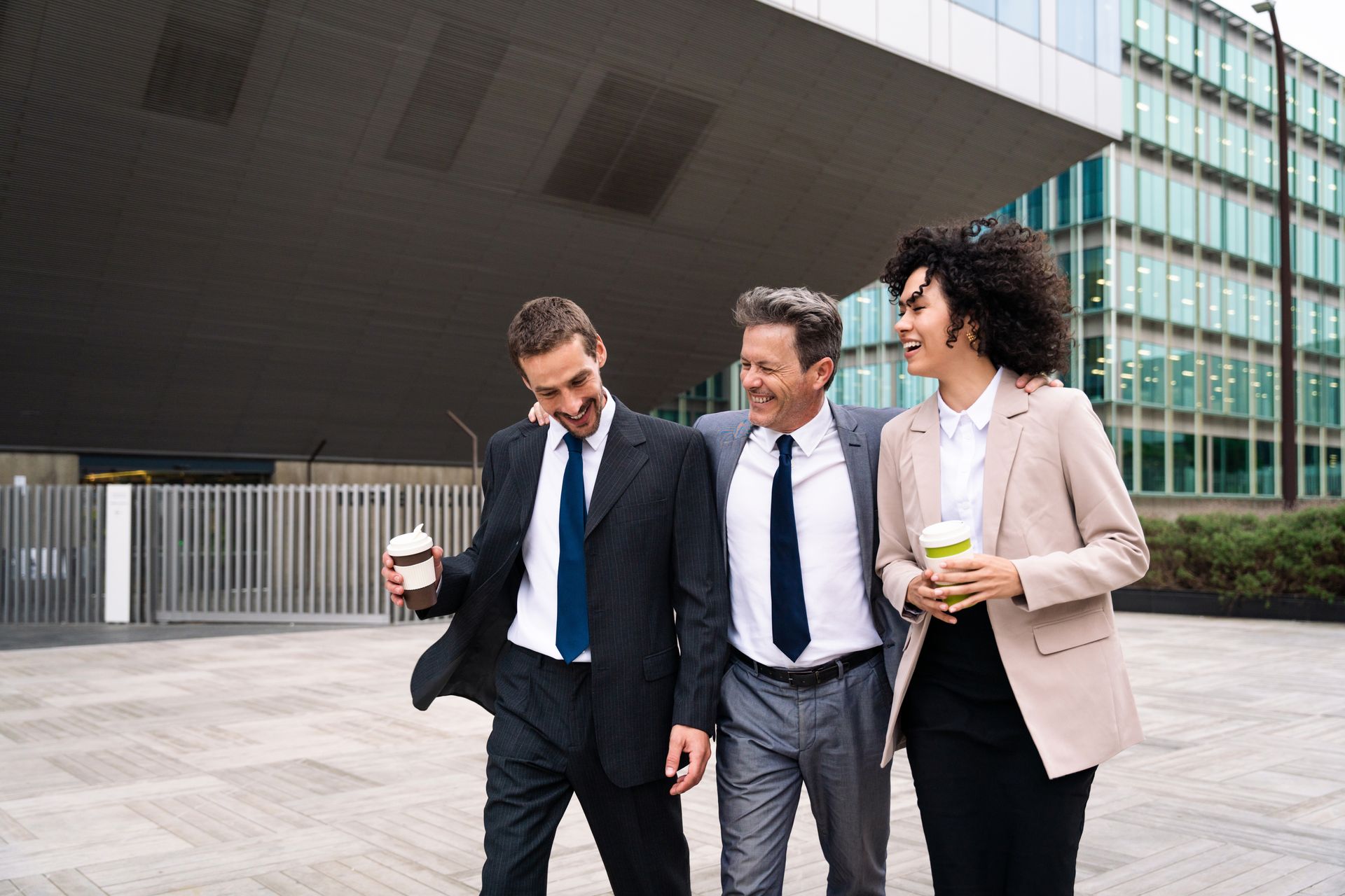 Three coworkers in business attire walking outside an office building, smiling and holding coffee cups.