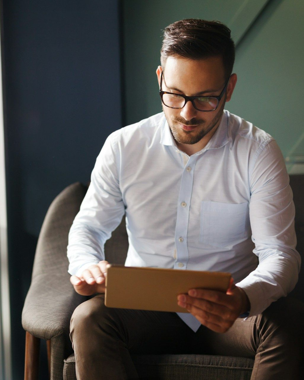 Man in a white shirt sitting in a chair, looking at a tablet in a softly lit room