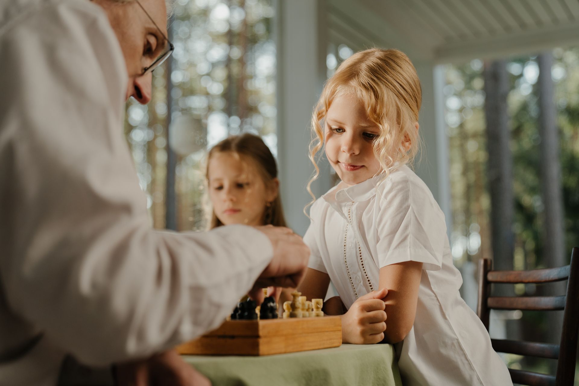 Adult and two children playing chess at a table in a bright room