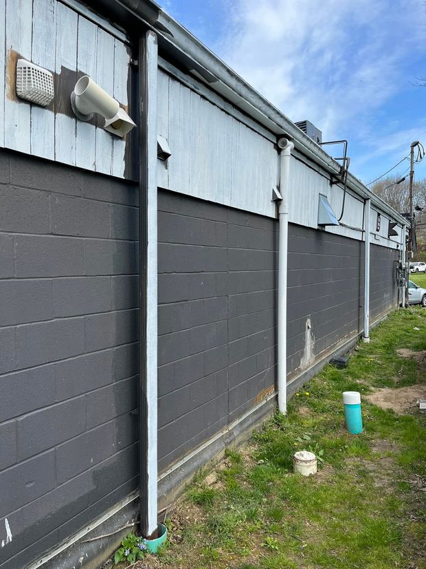 Side of a building with dark gray siding, white trim, and a few pipes against a blue sky.
