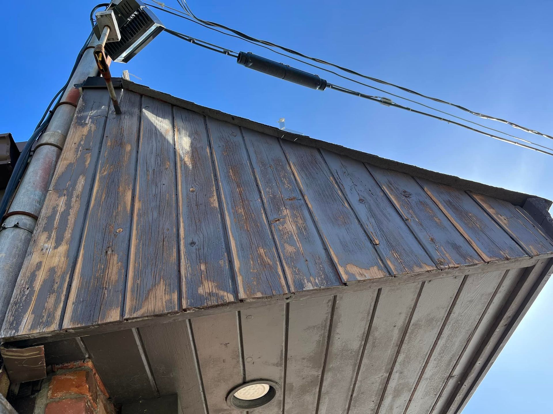 Low-angle view of a weathered, dark wooden awning under a clear, blue sky. Electrical wires and a pole are visible.