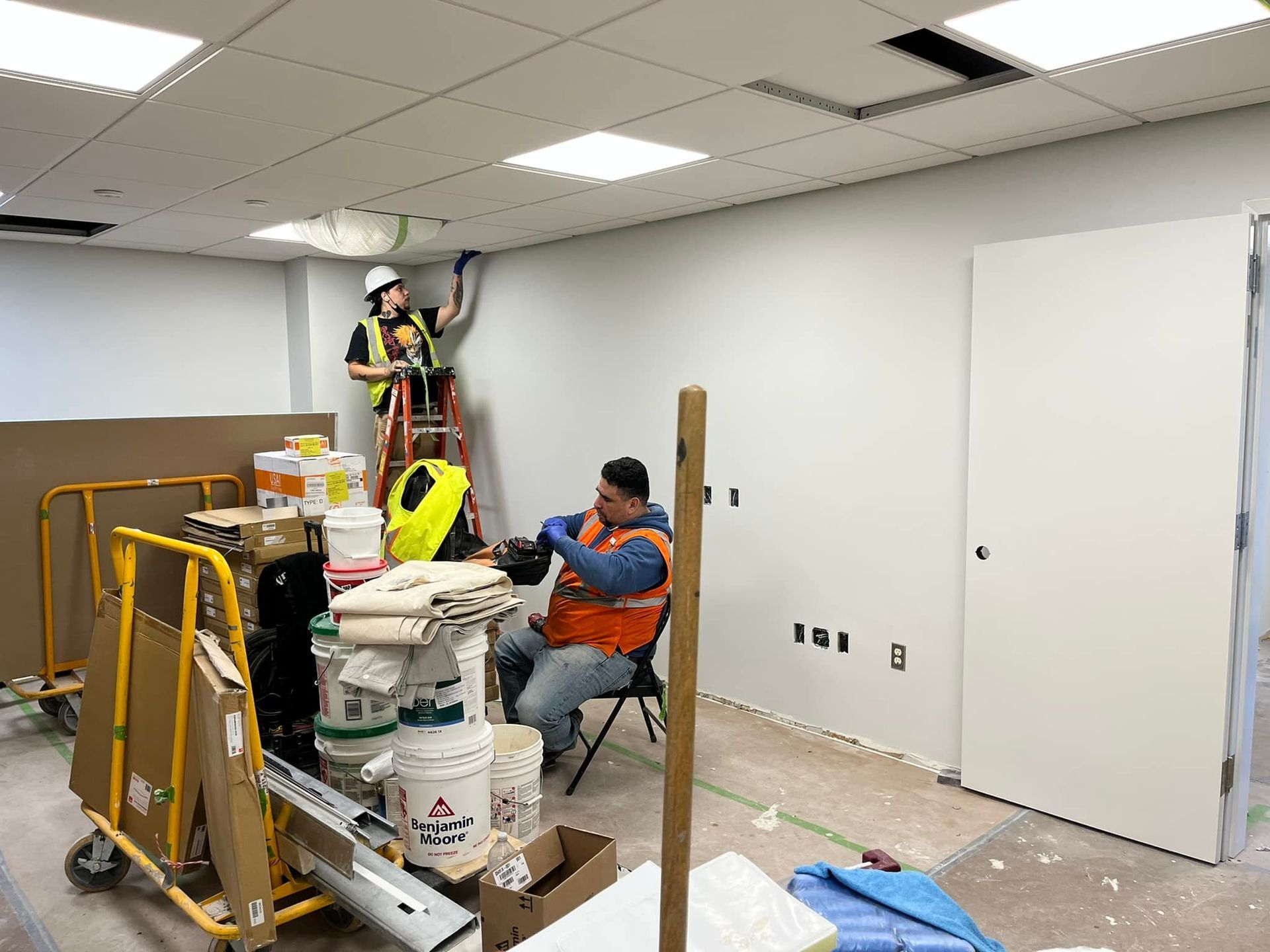 Two workers in hard hats install electrical wiring in a room under construction, one on a ladder, the other seated.