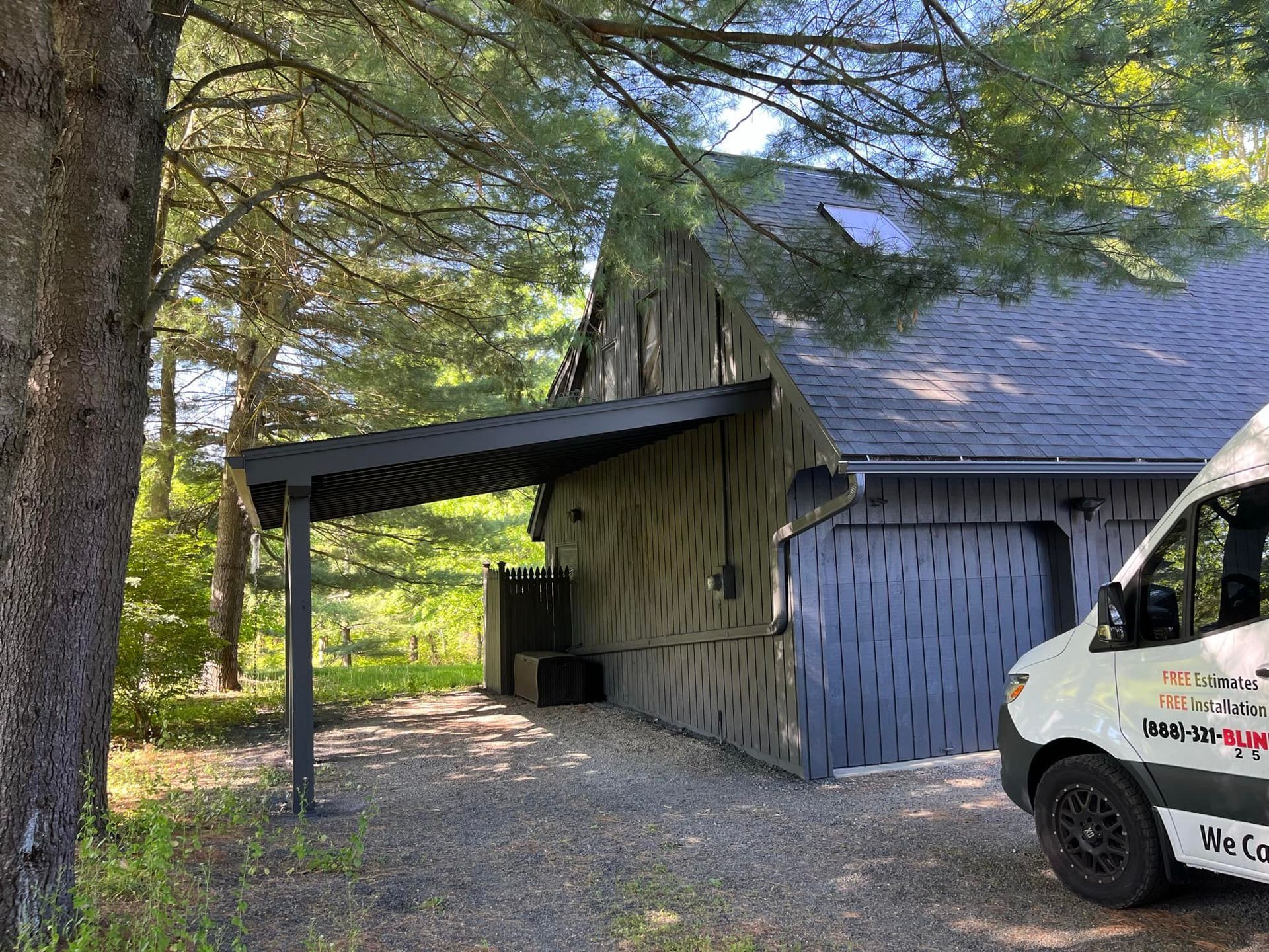 Building with dark siding, carport, gravel driveway, and a van. Sunlight and trees are in the background.