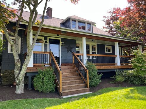 A house with a wraparound porch and steps, painted dark gray, with a star on the wall, and a green lawn.