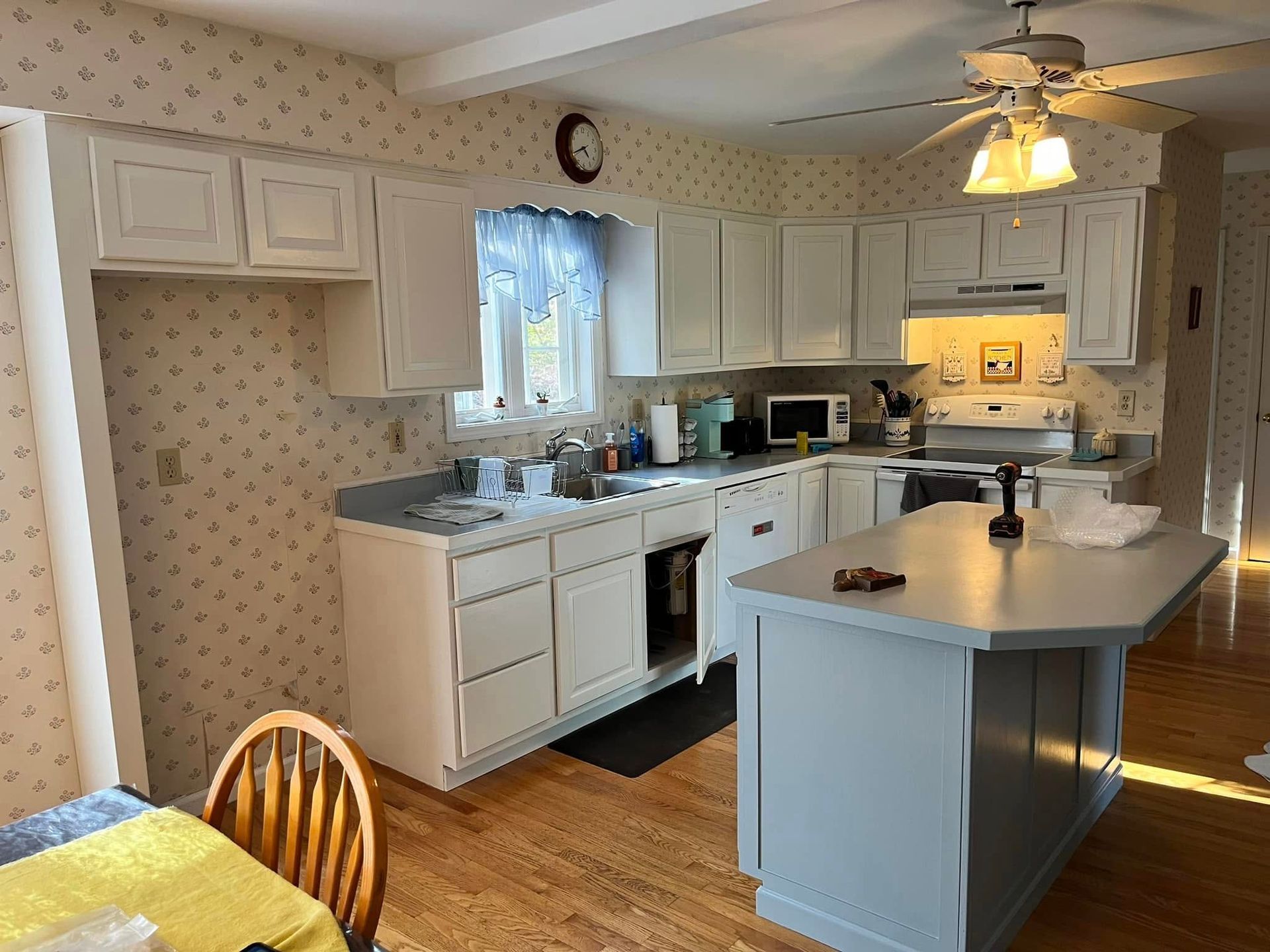 Kitchen with white cabinets, light blue countertops, wooden floor, island, wallpaper, and a window.