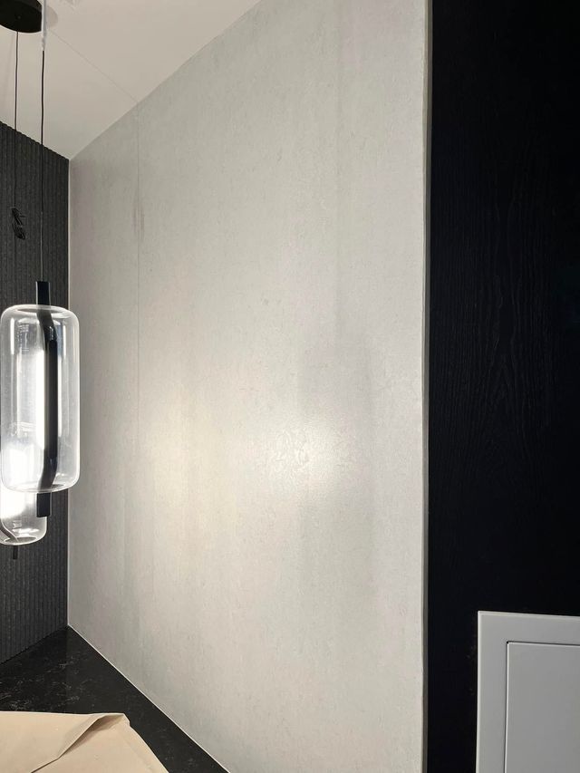 A modern interior featuring a textured light grey wall flanked by black panels, illuminated by a cylindrical pendant light.