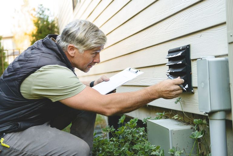 HVAC technician in overalls and gloves repairs wall-mounted air conditioner unit. Man examining a wall vent with a clipboard, kneeling outside a building.
