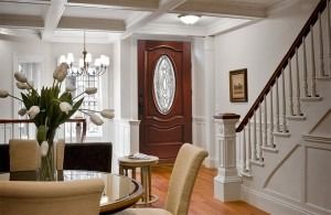 Bright foyer with staircase, oval front door, and dining table with flowers under white beamed ceiling