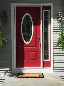 Red front door with oval glass panel and sidelights, framed by gray siding and a doormat.