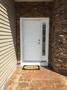 Front entry with white door, sidelight window, brick wall, and tan tiled porch with doormat.