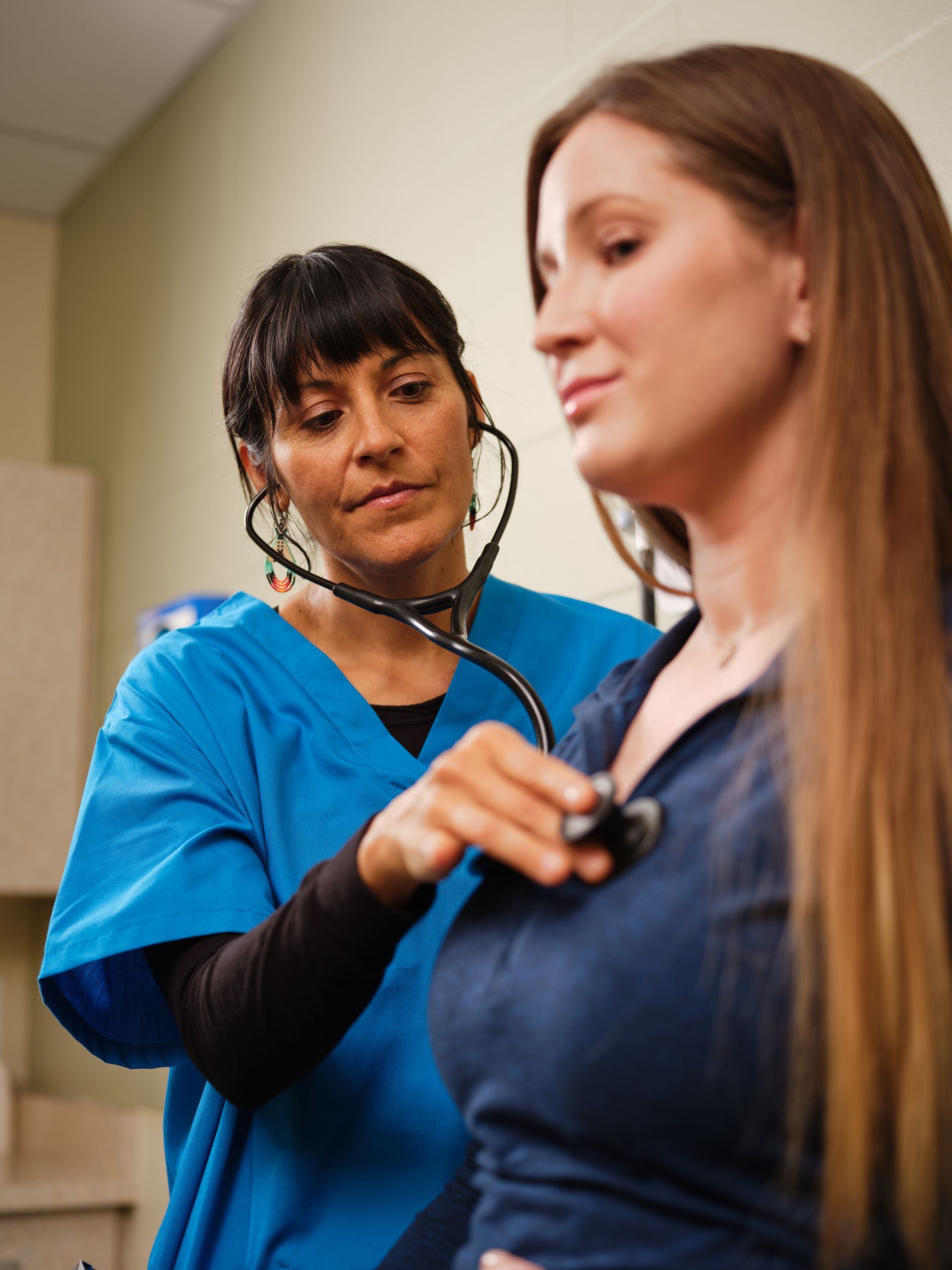 Nurse in blue scrubs listens with a stethoscope to a patient in a denim shirt.