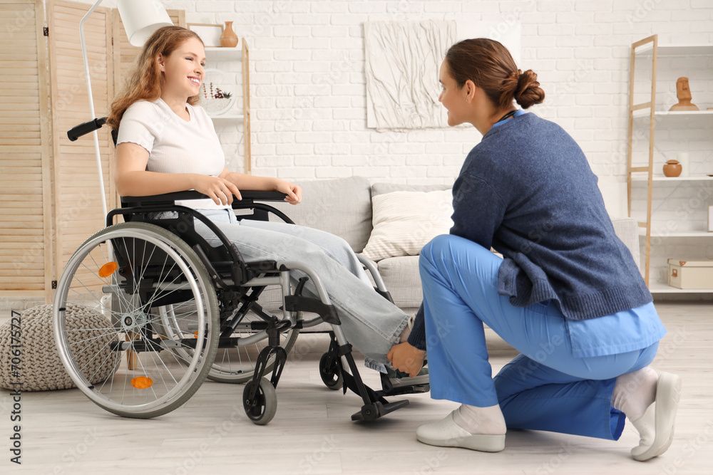 Nurse in blue scrubs listens with a stethoscope to a patient in a denim shirt. Caregiver kneels beside woman in wheelchair, adjusting footrest in a bright home living room.