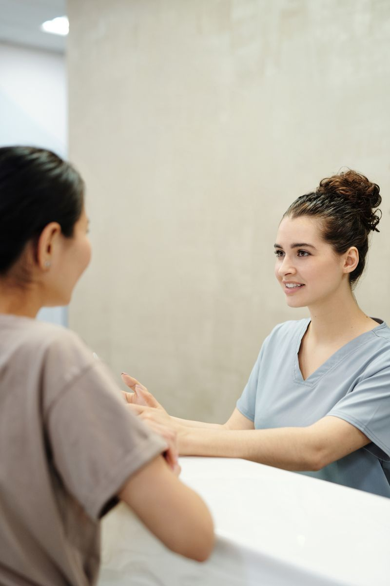 Nurse in blue scrubs listens with a stethoscope to a patient in a denim shirt. Caregiver kneels beside woman in wheelchair, adjusting footrest in a bright home living room.